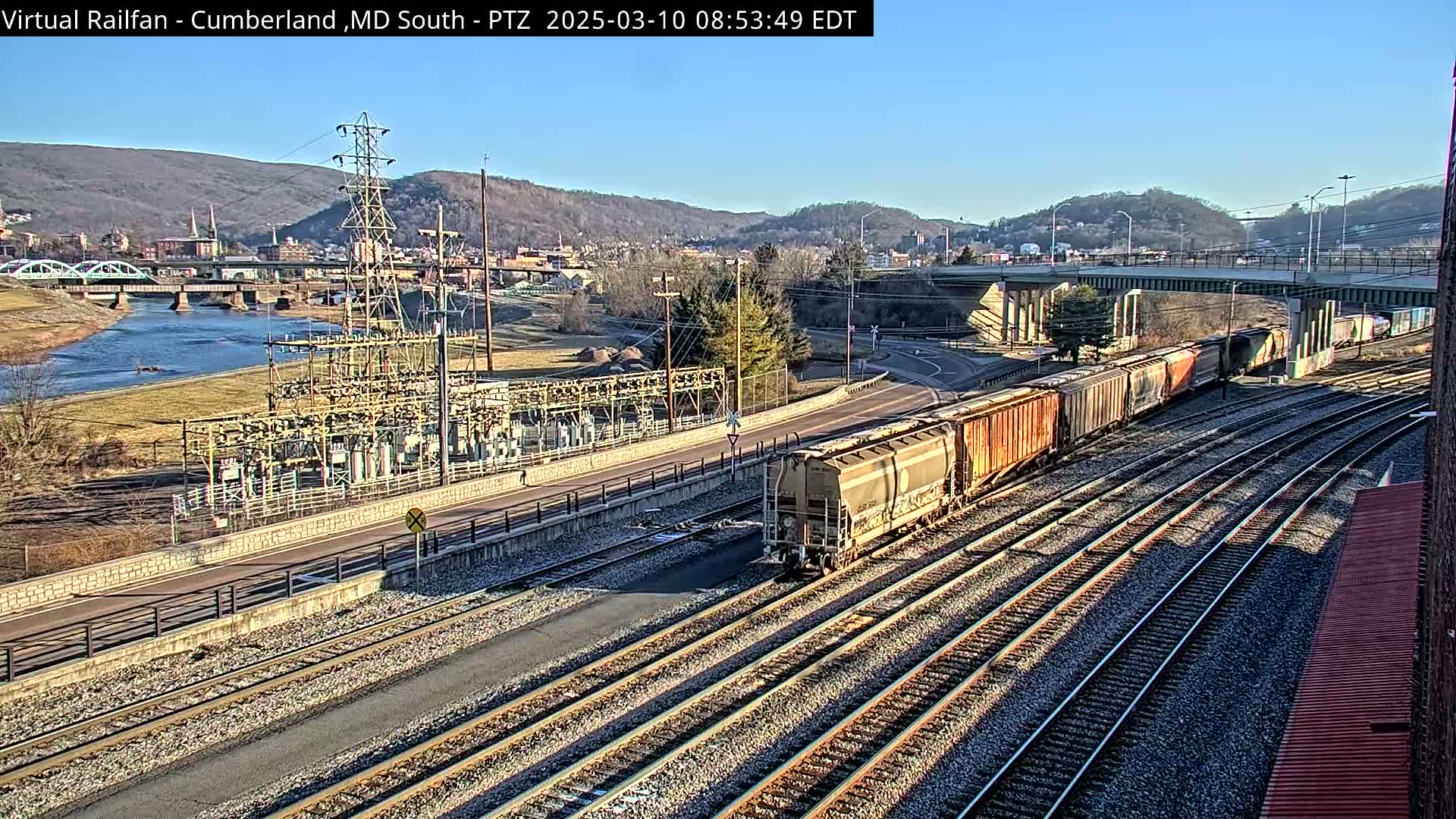 A long freight train is moving along multiple train tracks near a river and power station under a clear, sunny sky.