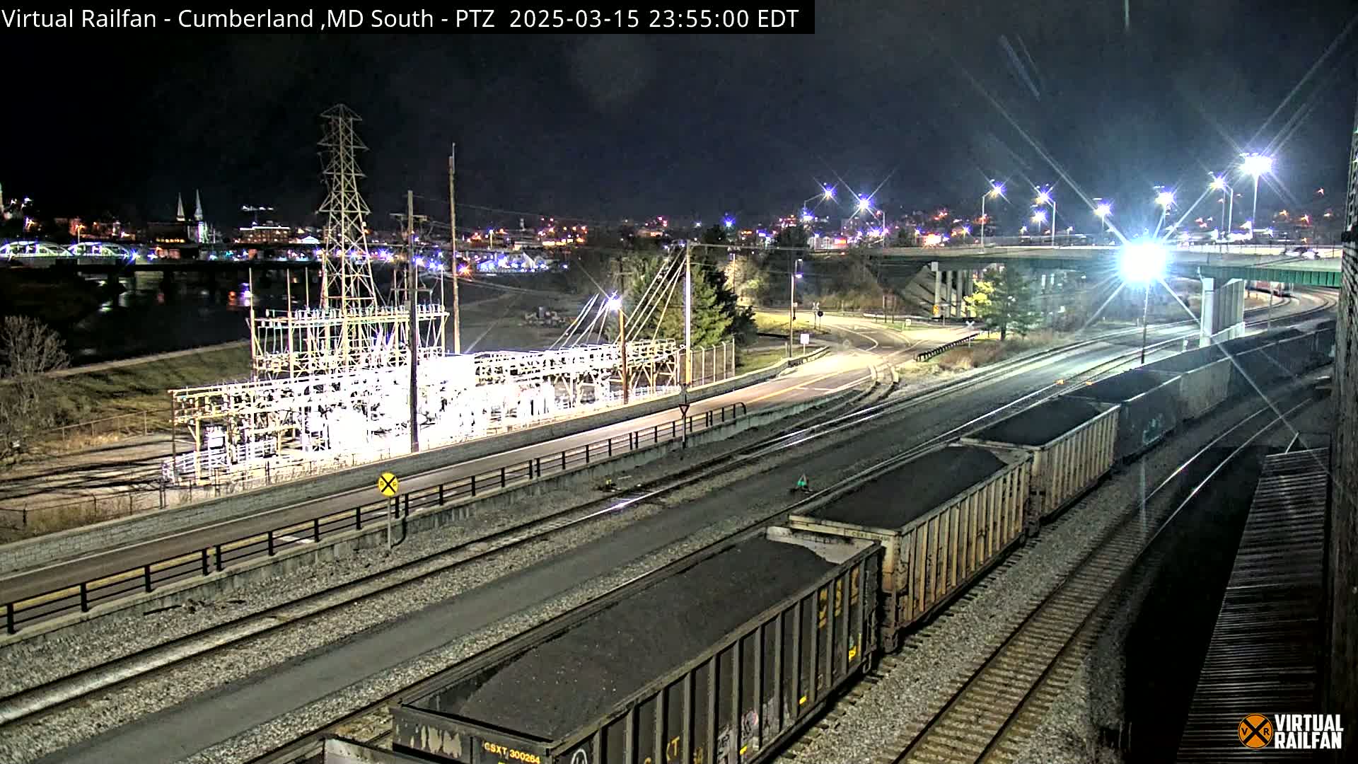 A nighttime view of a railway line with several coal cars, an electrical substation, and a river with city lights in the background under a clear sky.