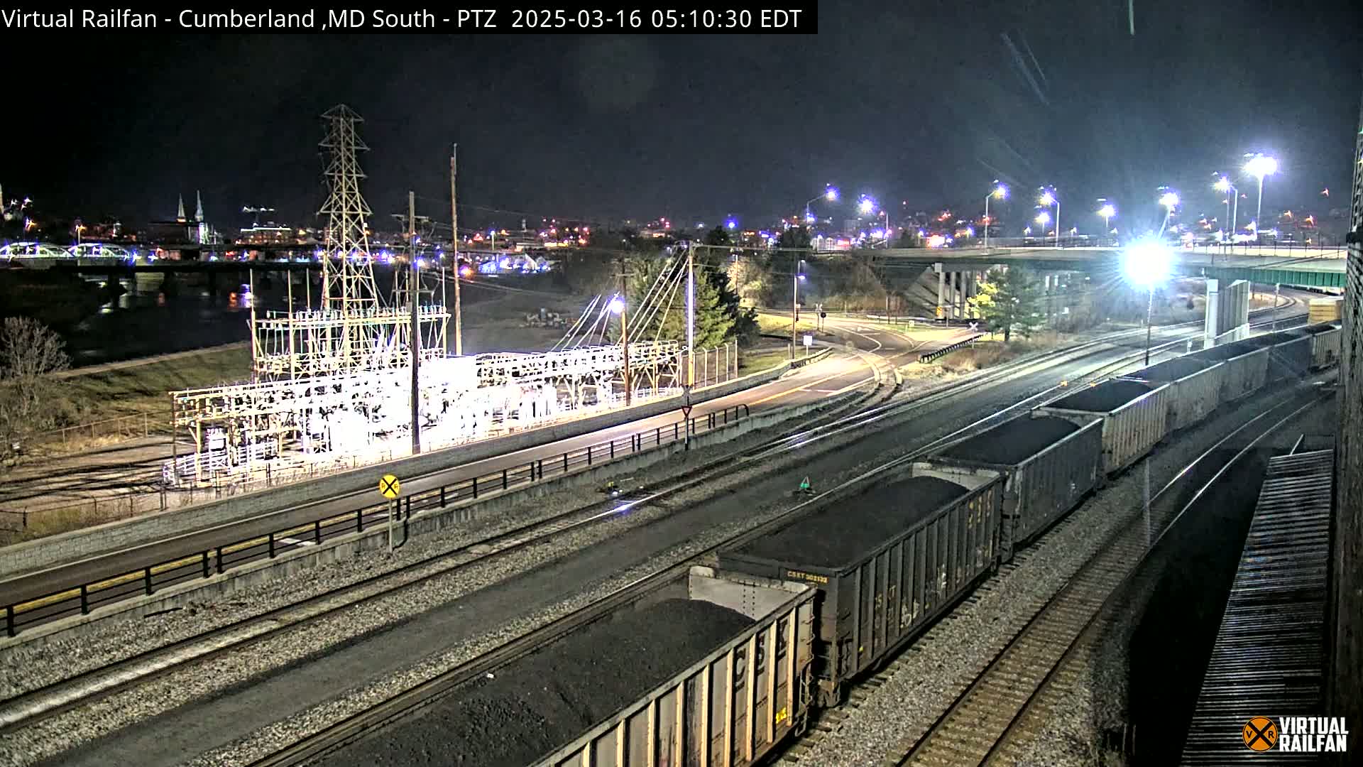 A nighttime view of a train carrying coal cars on railroad tracks near a power substation and a river, under well-lit conditions.