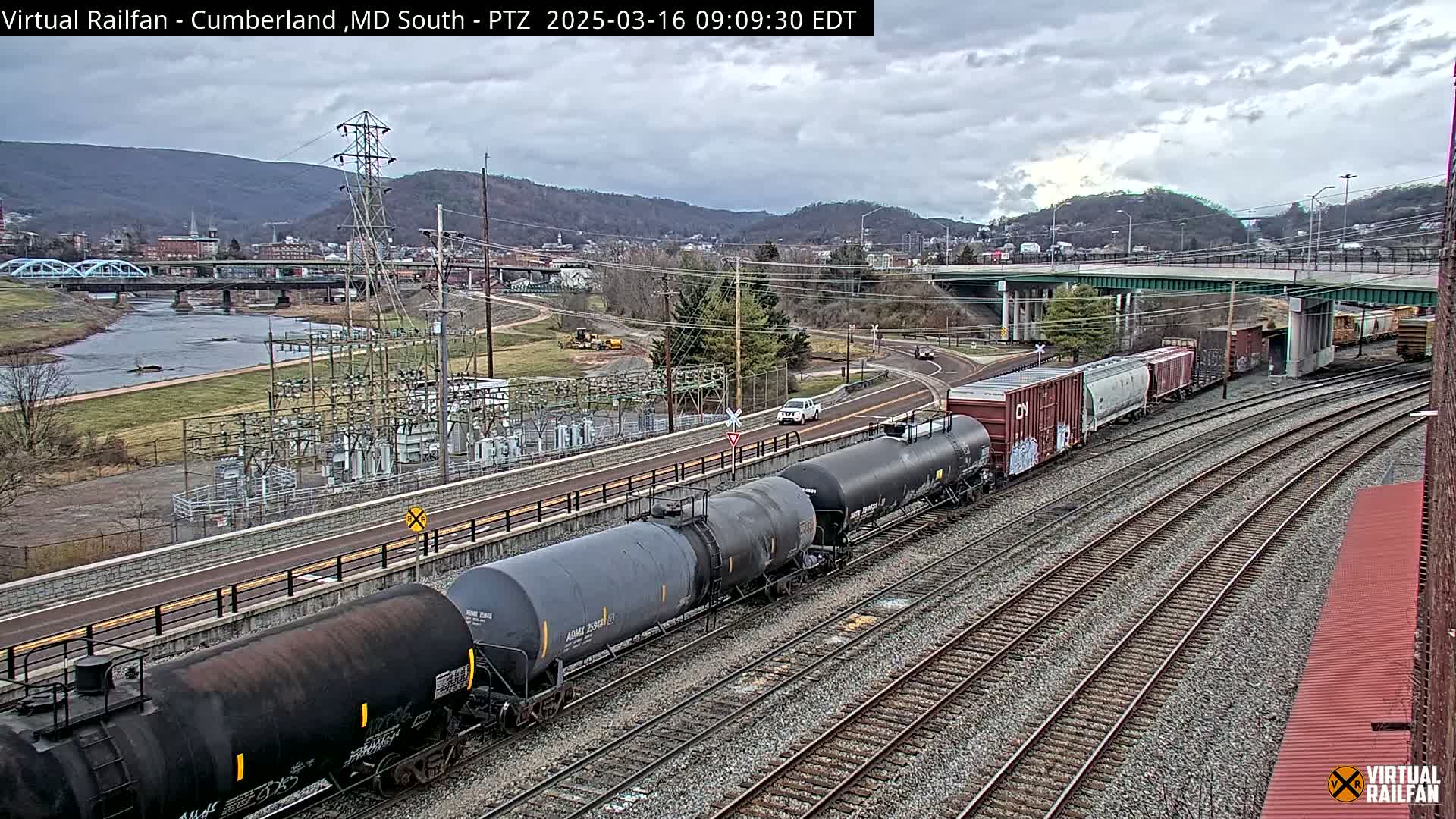 A long freight train of tanker cars and boxcars sits on a set of train tracks next to a river, under a cloudy sky, with a town and mountains in the background.