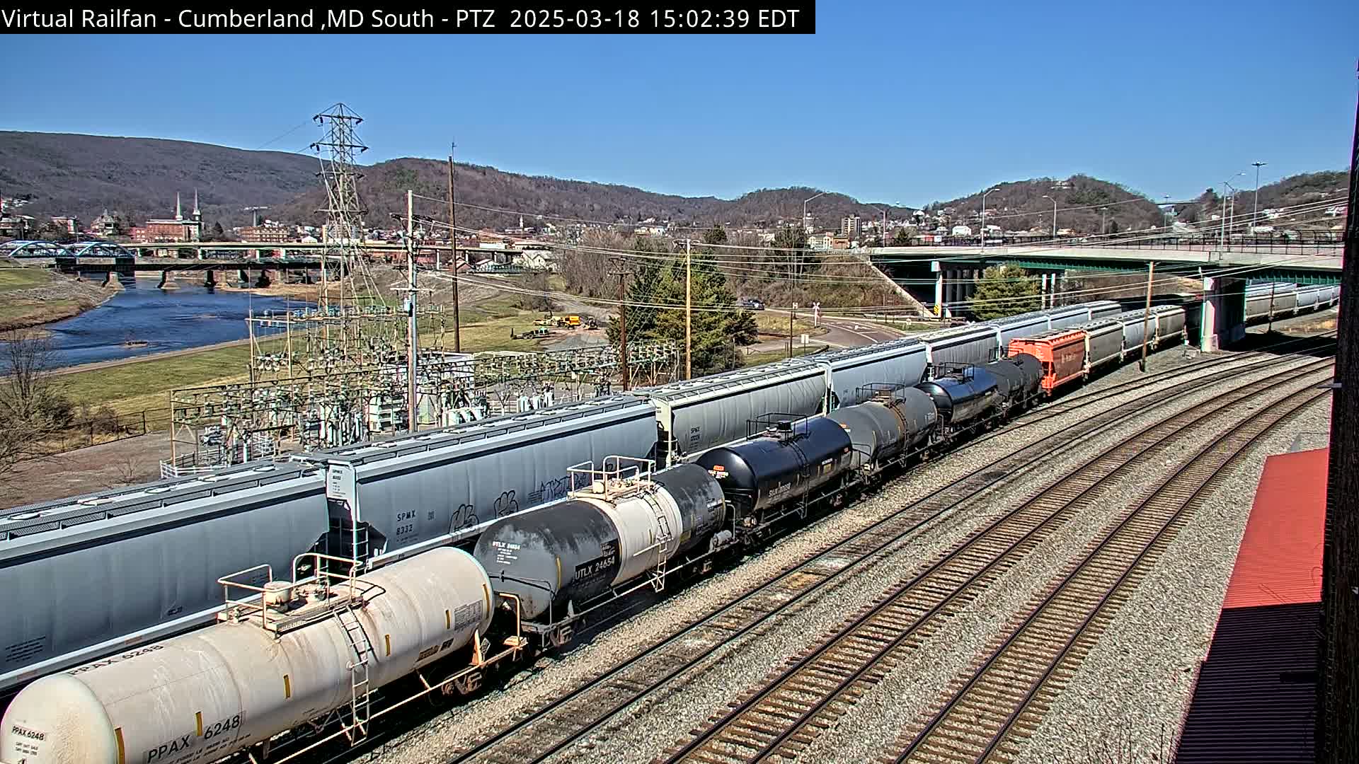 A long freight train carrying various cargo cars travels along a set of railway tracks under a clear, sunny sky, with a river and town visible in the background.