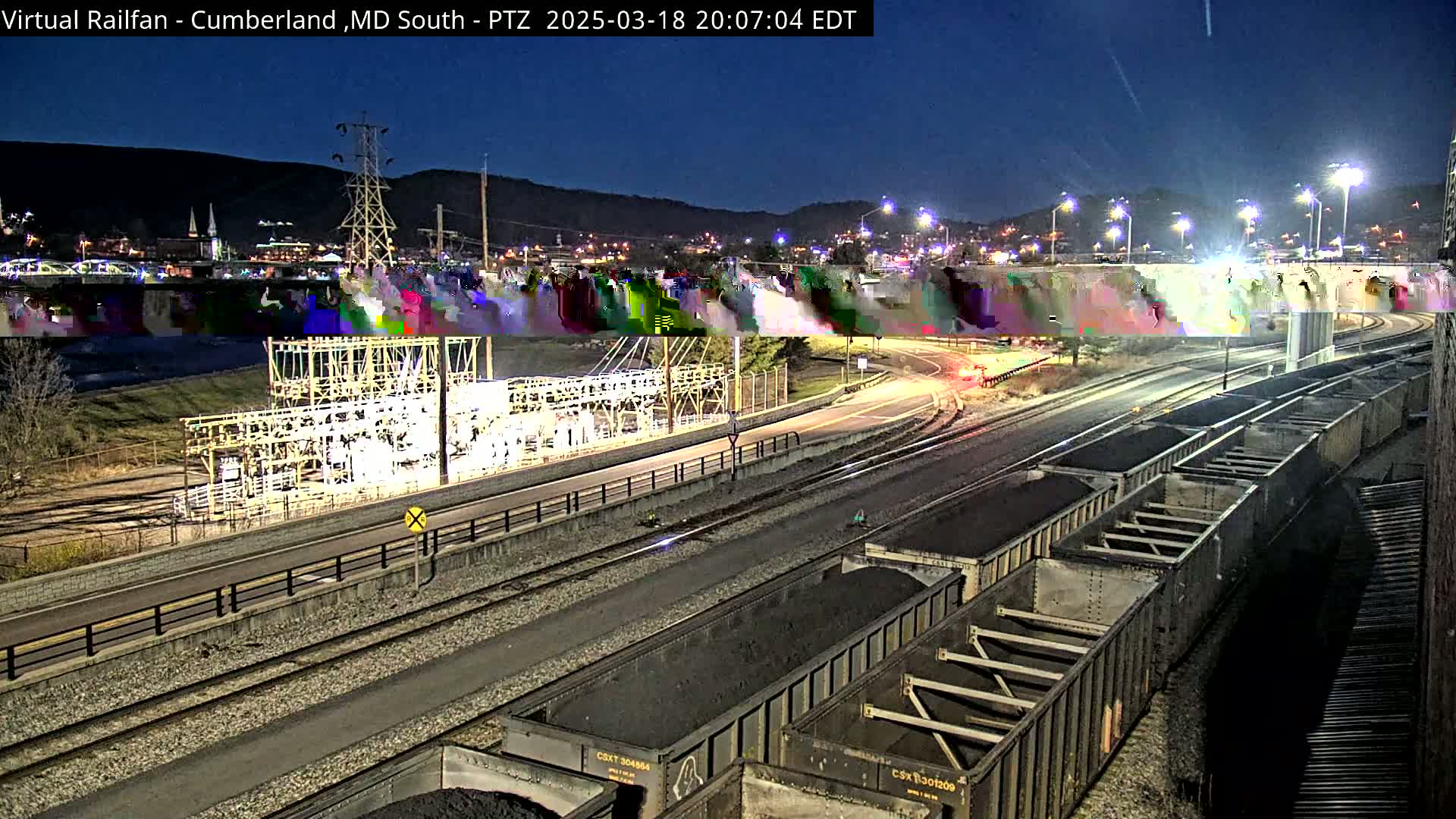 At night, under a dark sky, several coal cars sit on railroad tracks next to a roadway and an electrical substation, with a town visible in the background.
