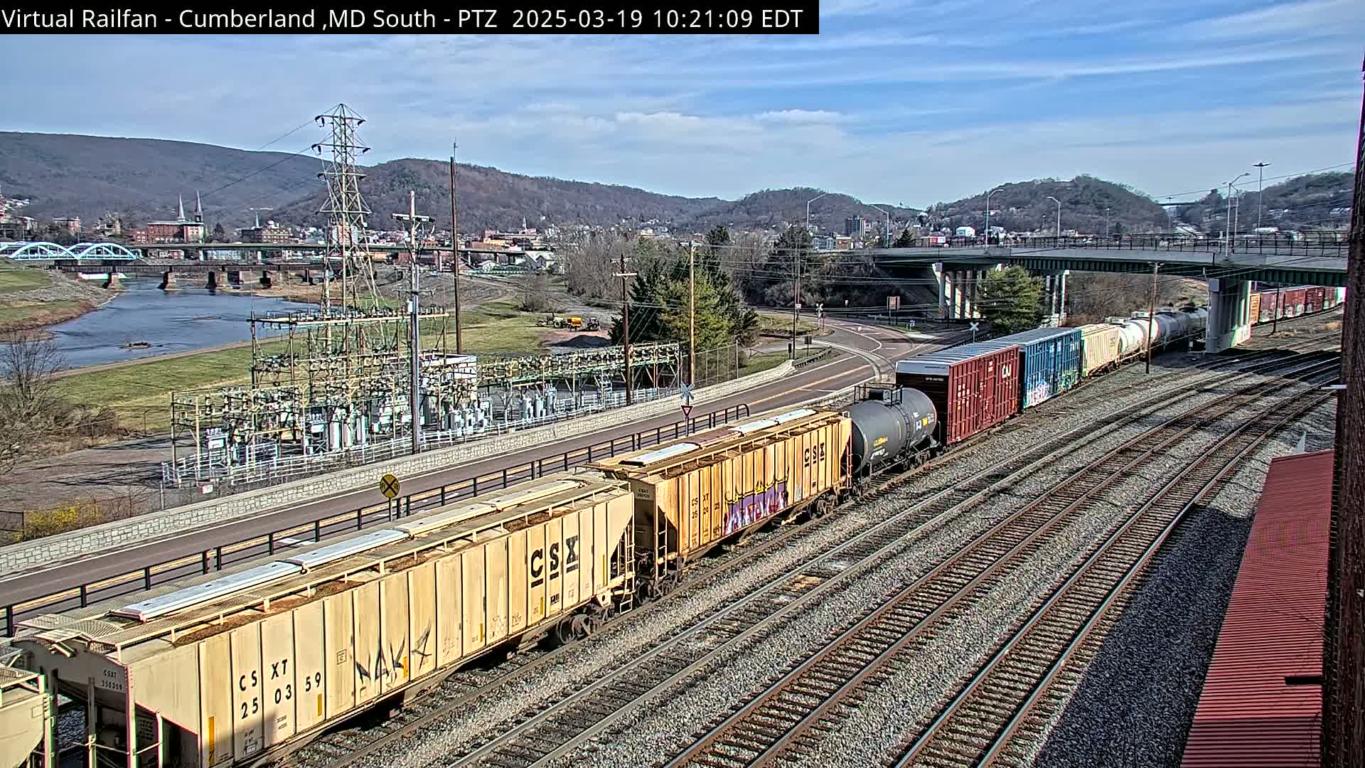 A freight train carrying various cargo cars travels along a set of railroad tracks, with a river, a bridge, and a town visible in the background under a partly sunny sky.