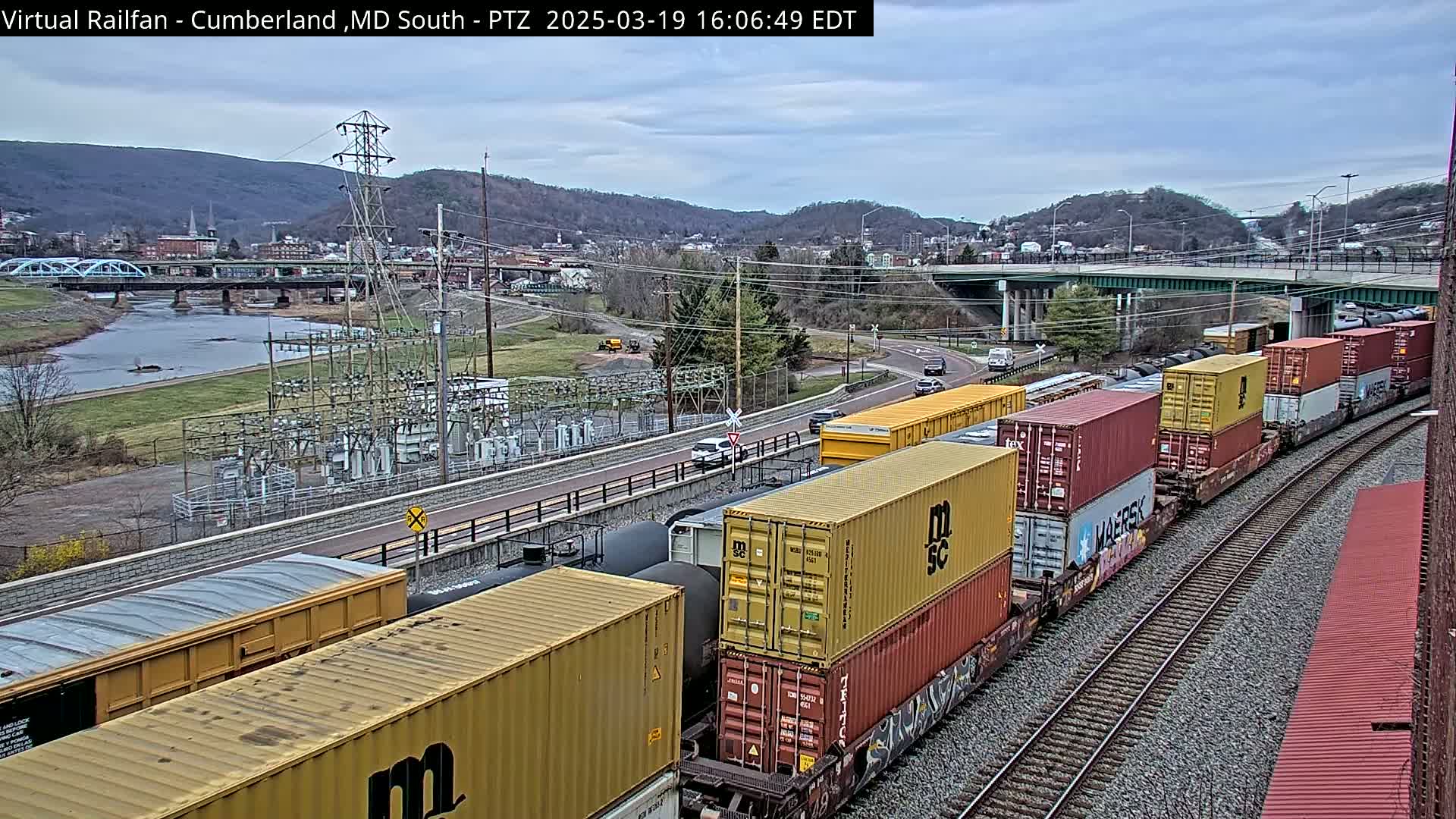 A long freight train carrying multiple shipping containers travels along railroad tracks next to a river and a town under an overcast sky.