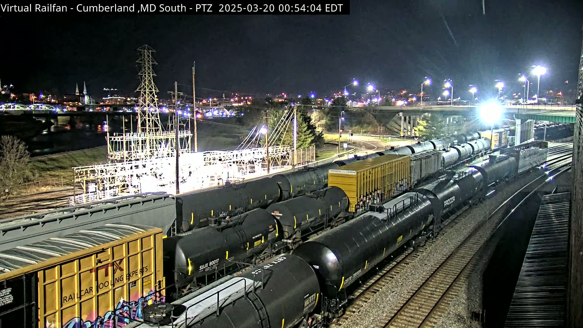 A long freight train of tanker cars is parked at night near a power substation and a river, under well-lit conditions.