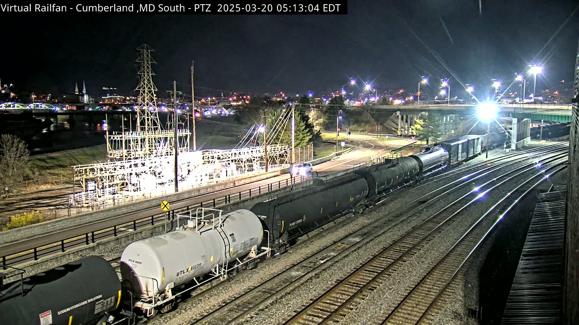 At night, a long freight train of tanker cars passes an electrical substation next to a roadway and a bridge, under bright artificial lights.