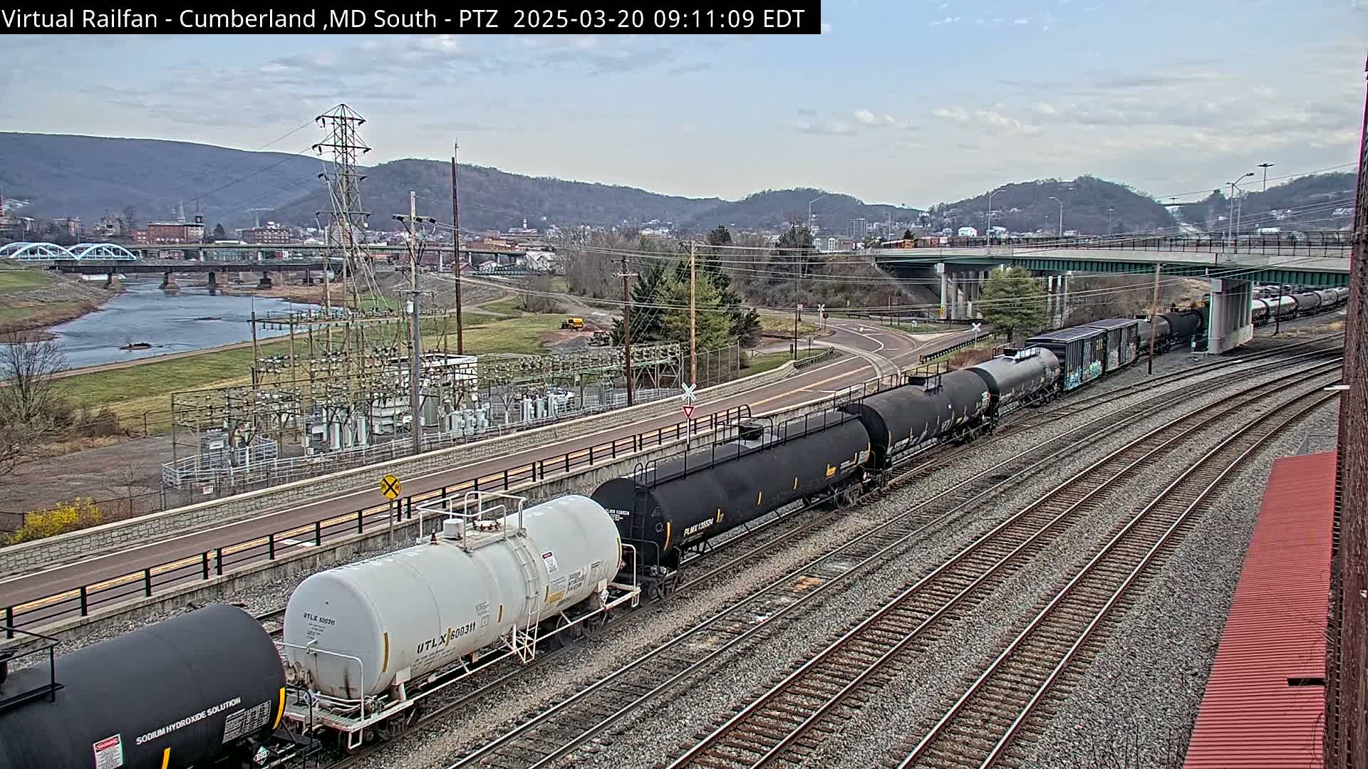 A long freight train of tanker cars travels along railroad tracks beside a river, under a mostly clear sky, with a town and hills in the background.