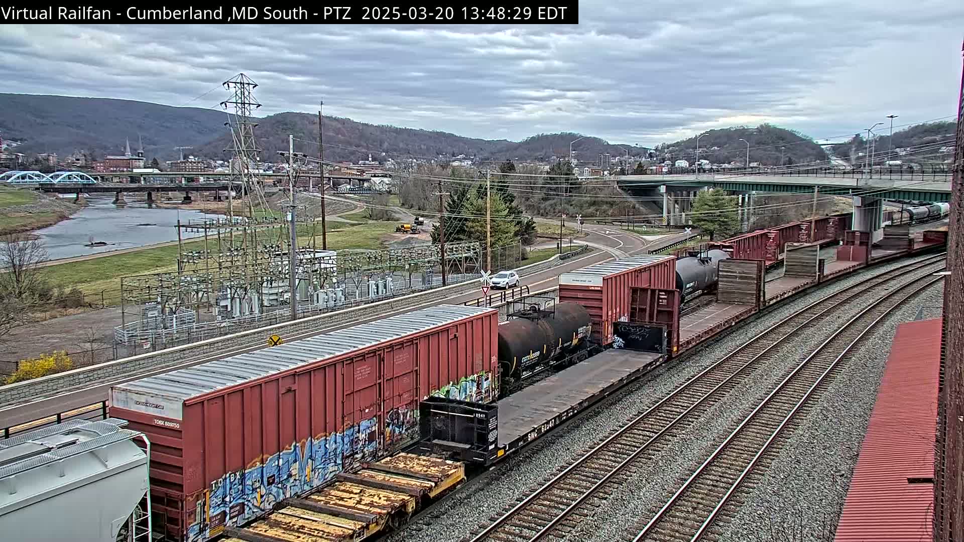 A long freight train with graffiti-covered cars curves along railroad tracks under an overcast sky, passing by a river, a bridge, and a power station nestled against a hillside.