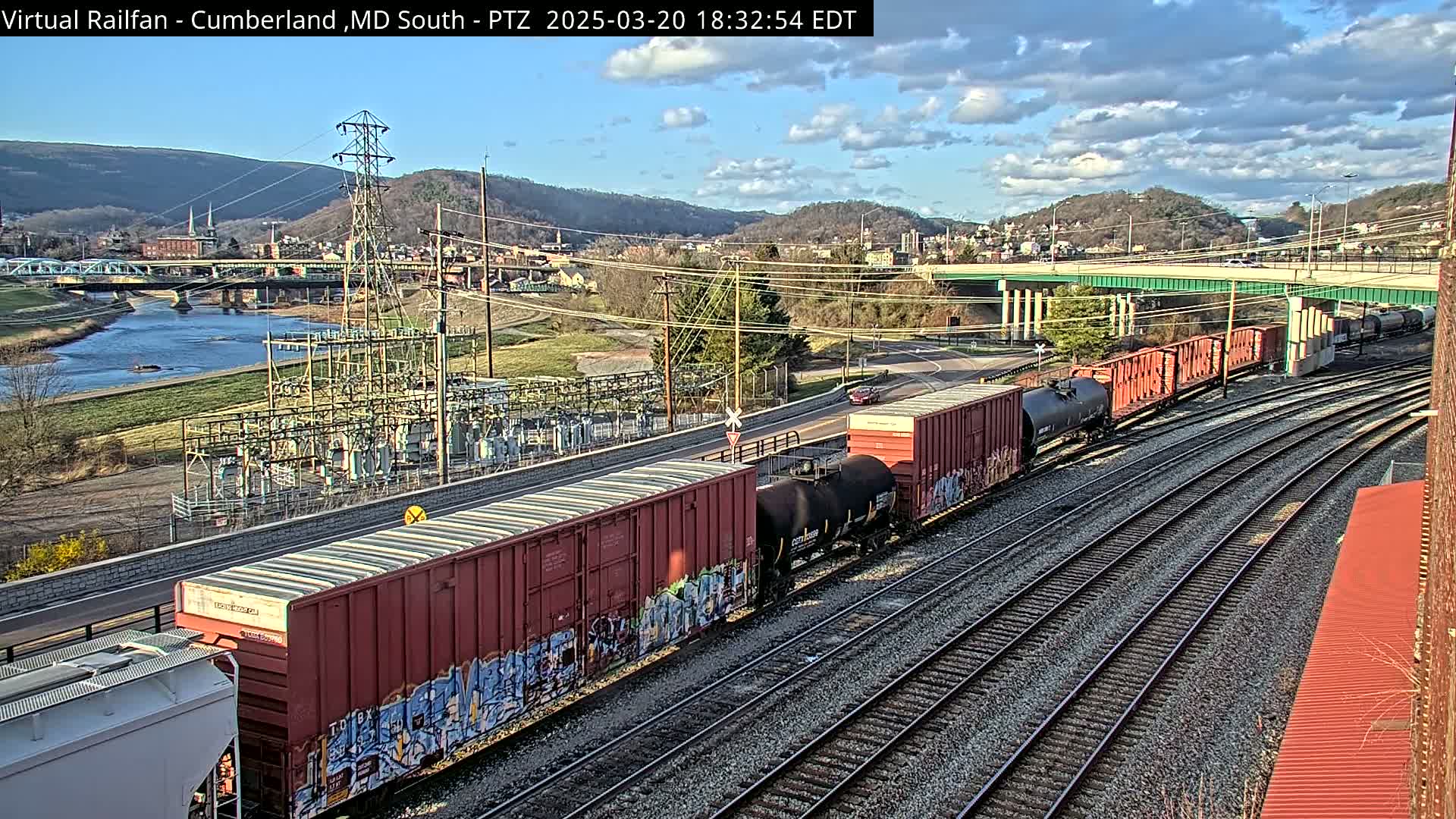 A long freight train with graffiti-covered cars sits on a set of tracks next to a river and a small town under a partly cloudy sky.