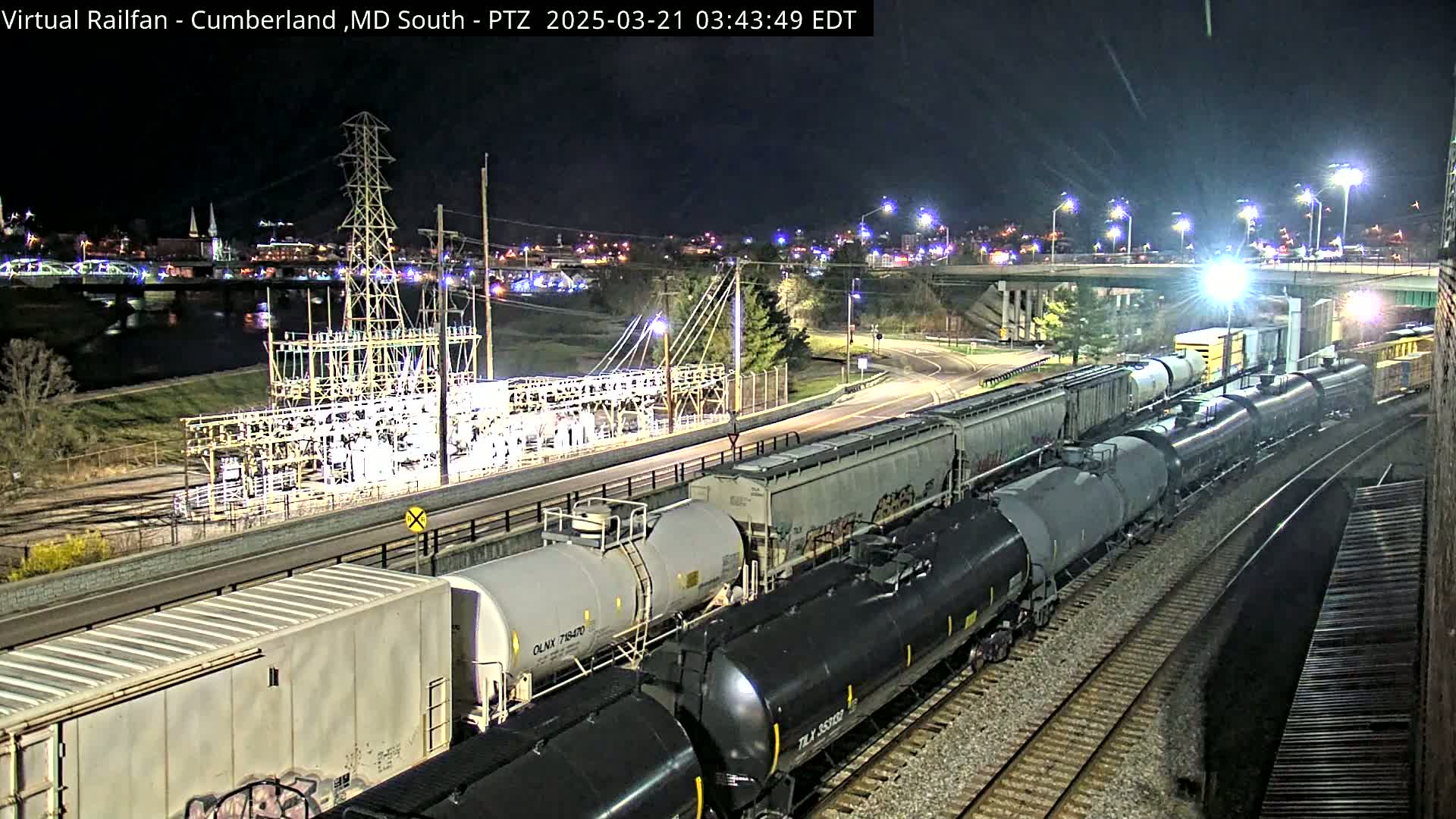At night, a long freight train of tanker cars and boxcars travels along railroad tracks near a power substation and a river, under bright streetlights.
