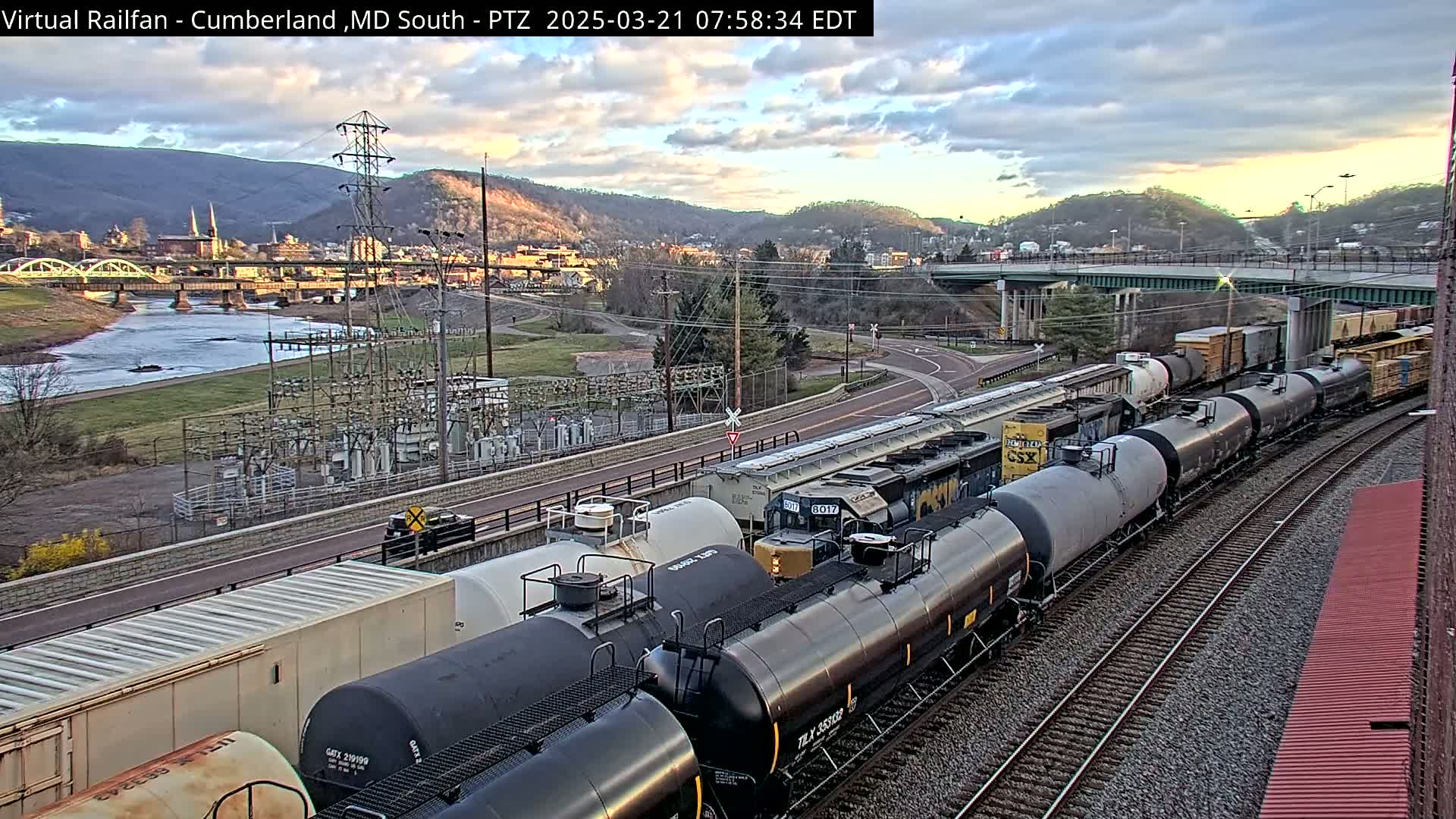 A long freight train carrying tanker cars is seen on a set of railroad tracks next to a river and a town, under a partly cloudy sky.