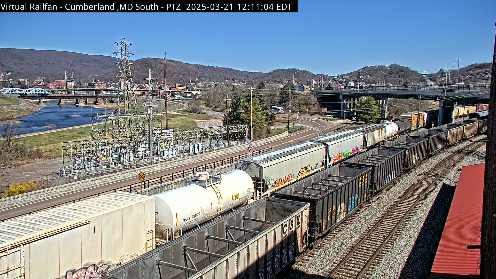 A long freight train, some cars graffitied, passes by a river, a bridge, and a town under a clear blue sky.