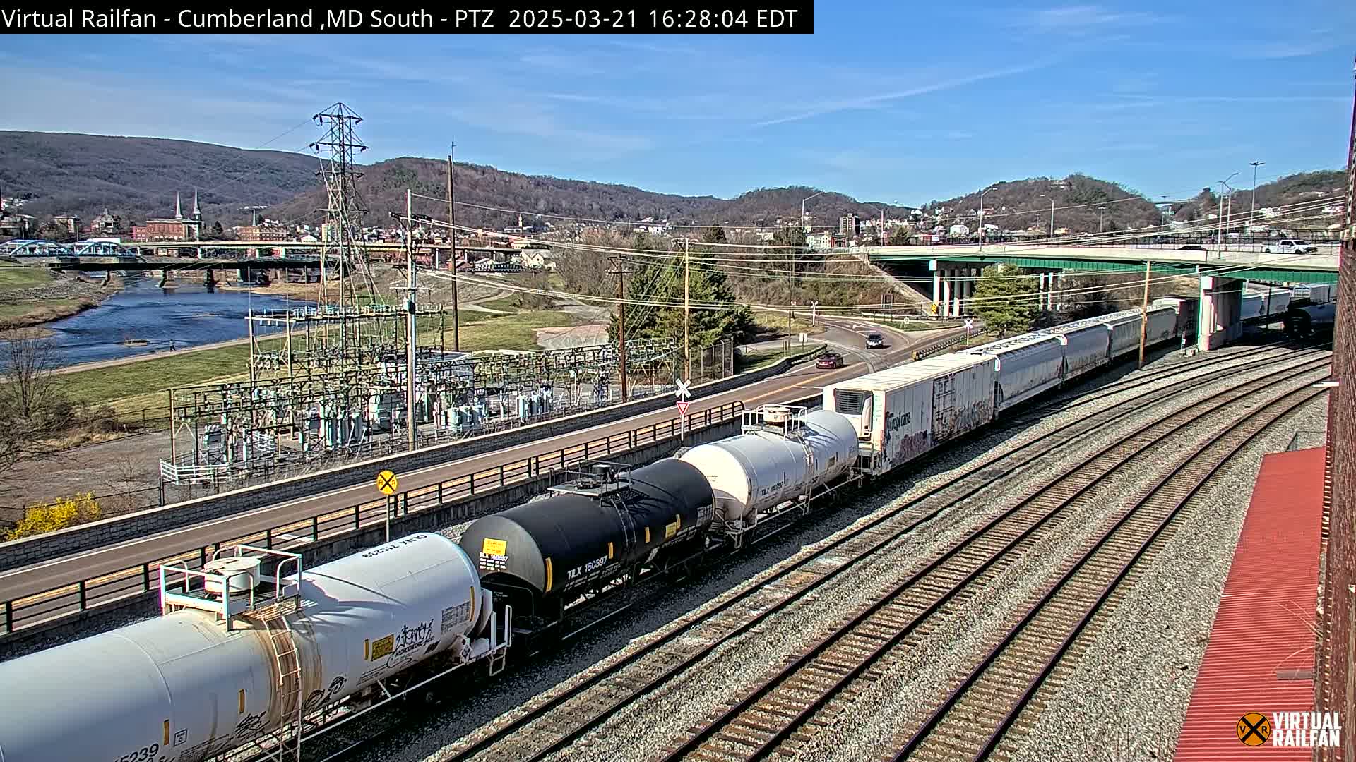 A long freight train carrying tanker cars and boxcars is traveling along railroad tracks on a sunny day, with a river, a bridge, and a town visible in the background.