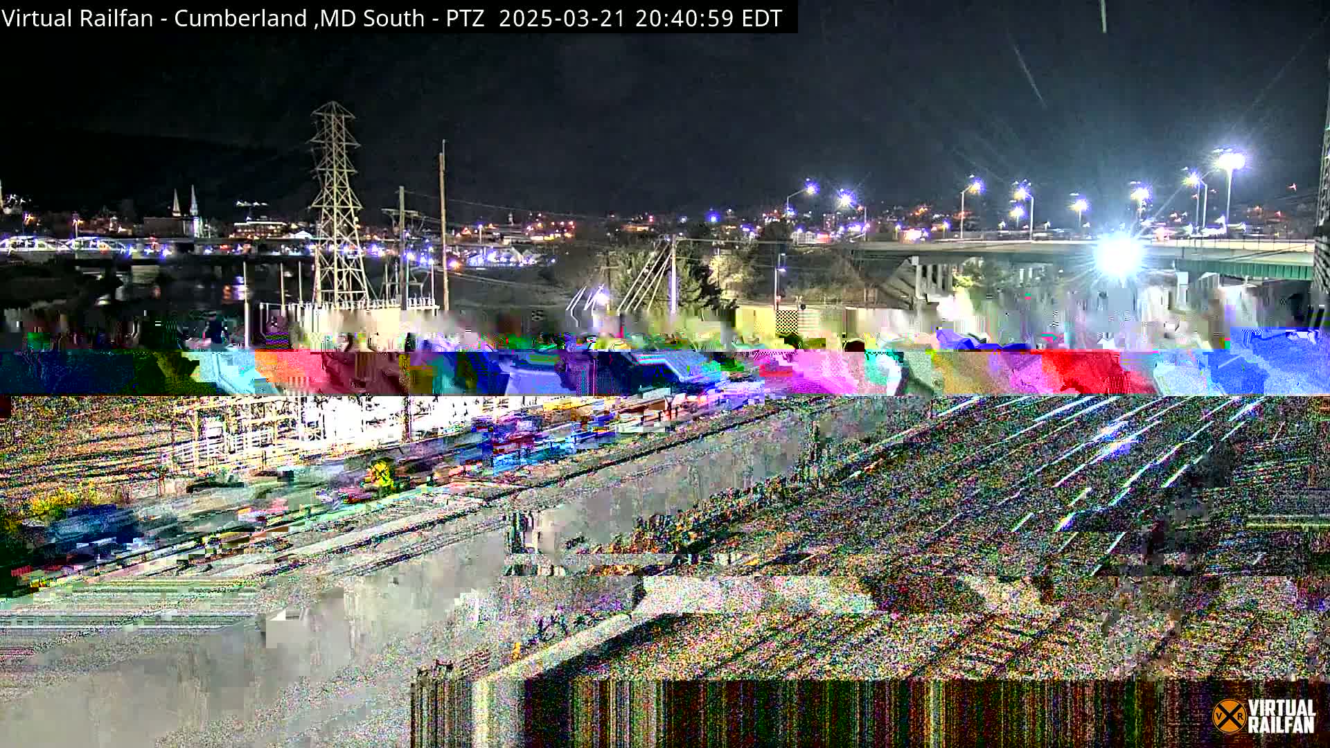 A nighttime view of a rail yard with a bridge and city lights in the background, showing significant image corruption.
