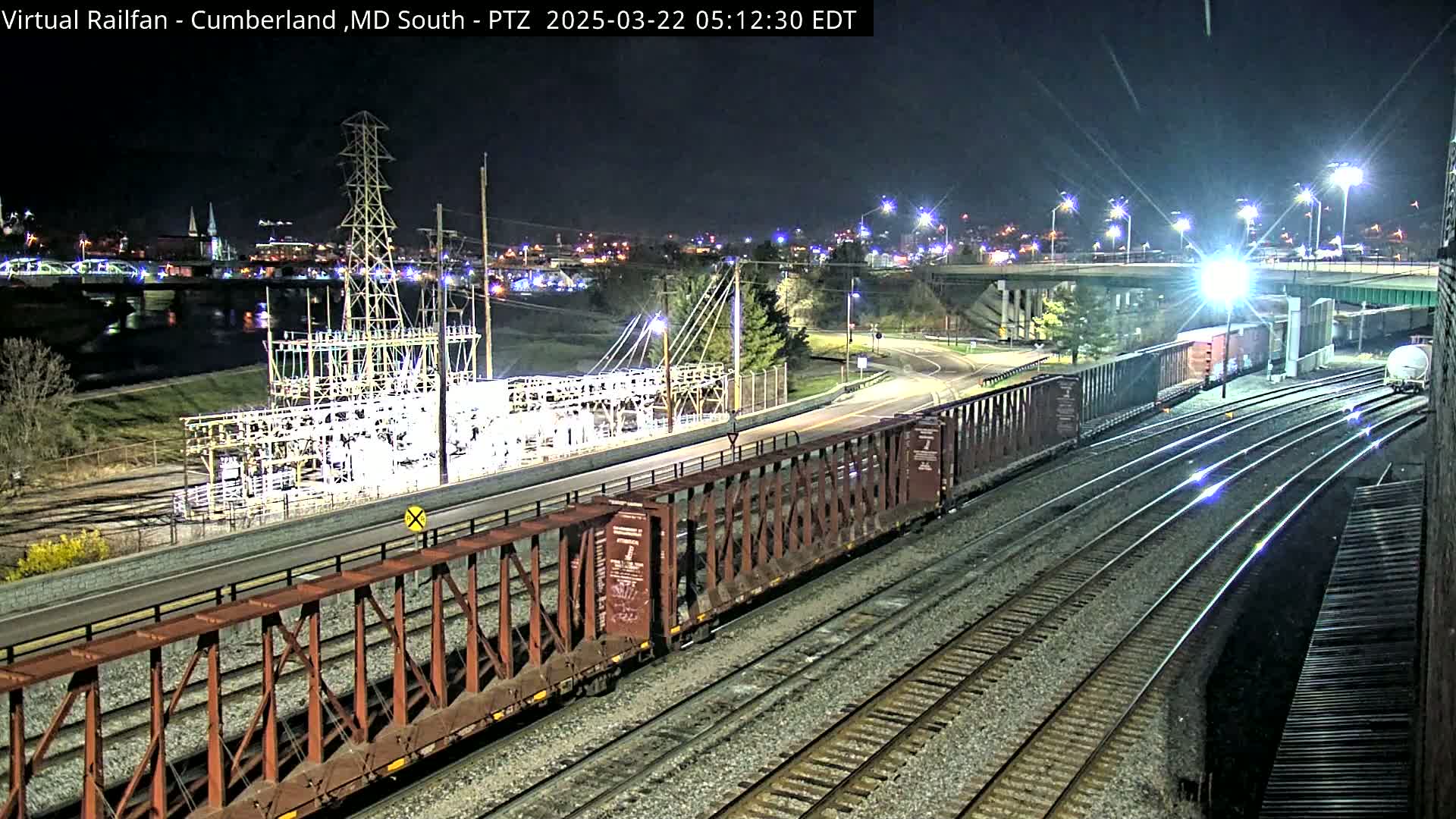 At night, a long freight train sits on railroad tracks next to a road and an electrical substation, all illuminated by streetlights.