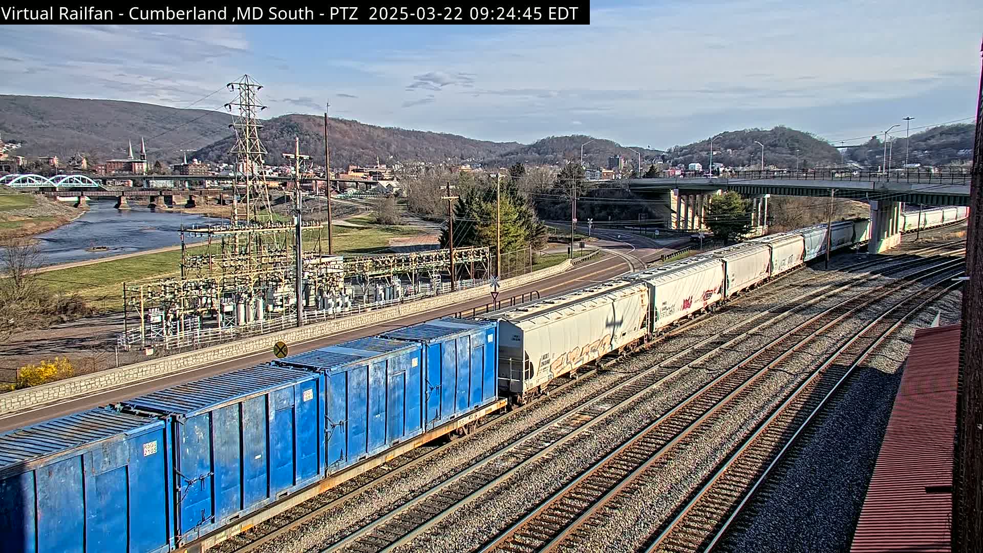 A long freight train carrying blue and white containers passes through a valley town under a bridge on a partly sunny day.