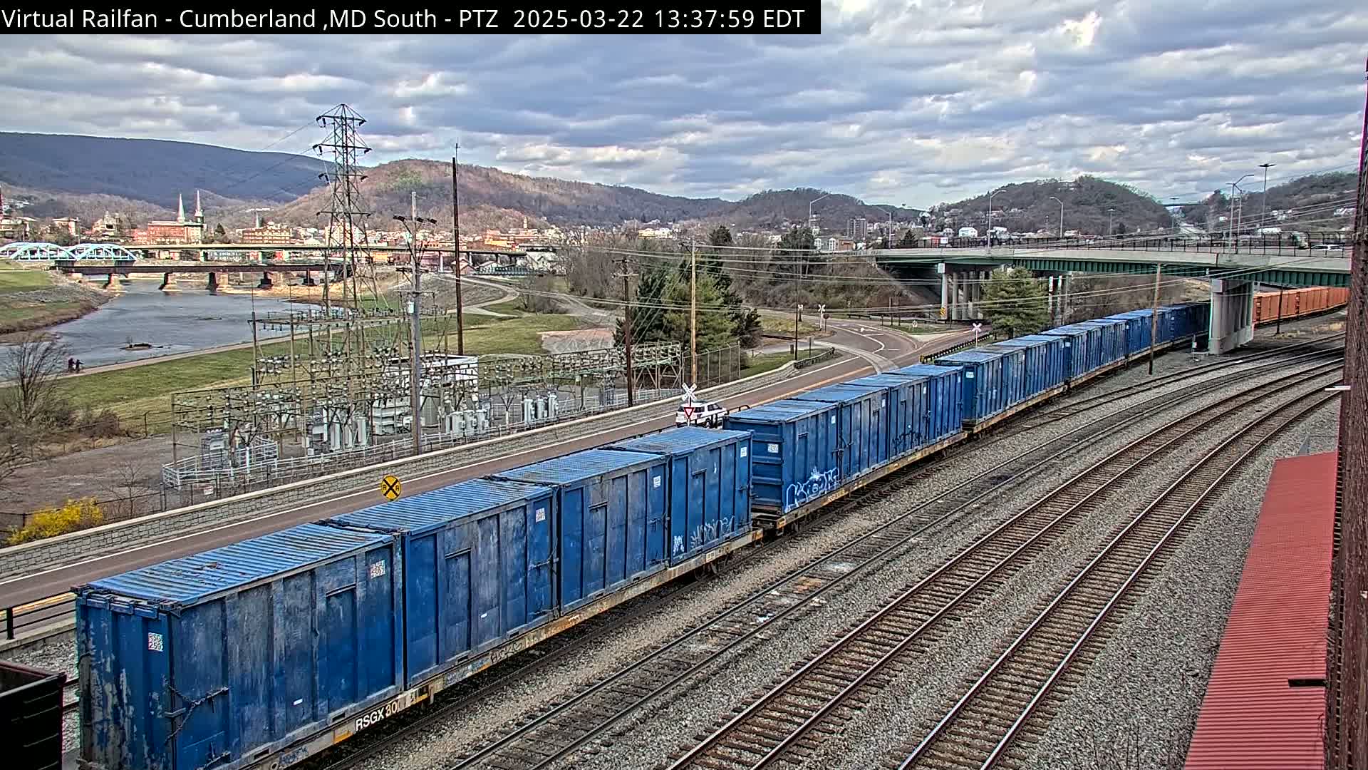 A long freight train carrying blue shipping containers travels along train tracks, under a partly cloudy sky, past a river and a town nestled in a valley surrounded by hills.