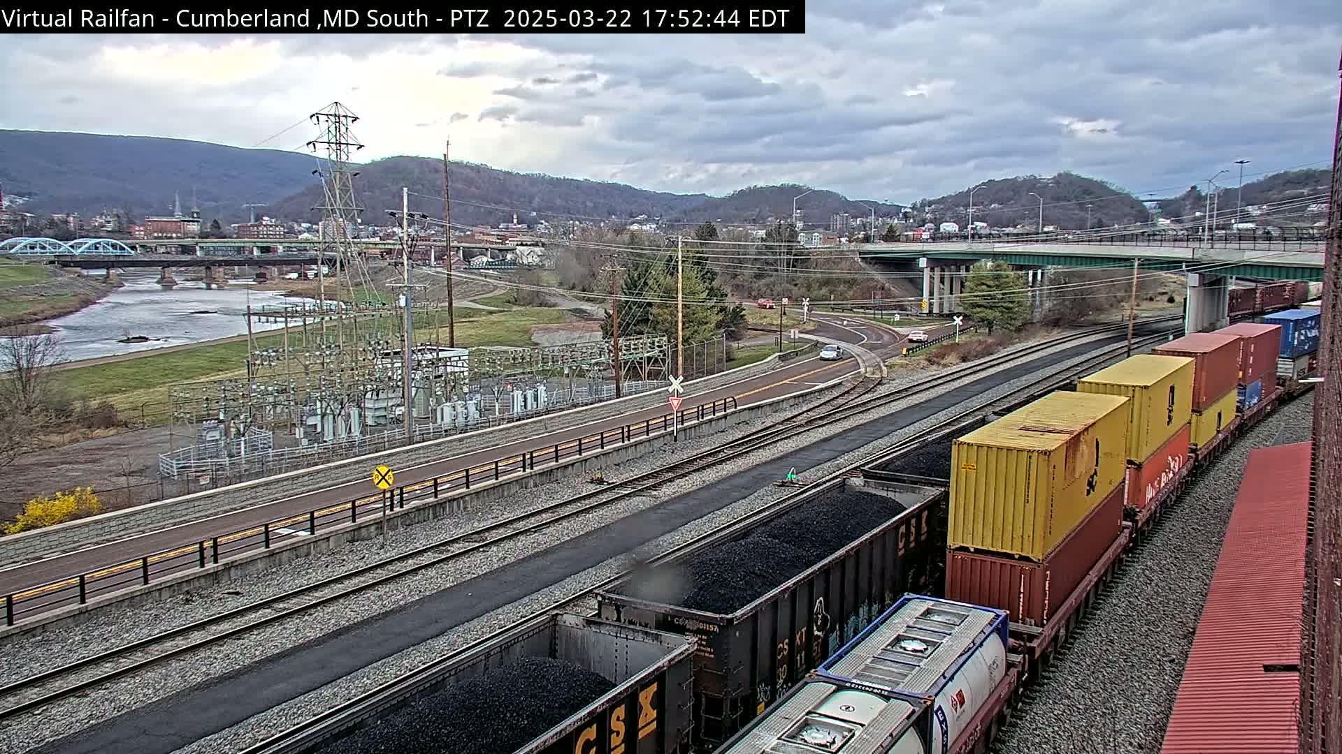 A train carrying coal and shipping containers sits on railroad tracks next to a river, under a cloudy sky, with a power station and a bridge visible in the background.