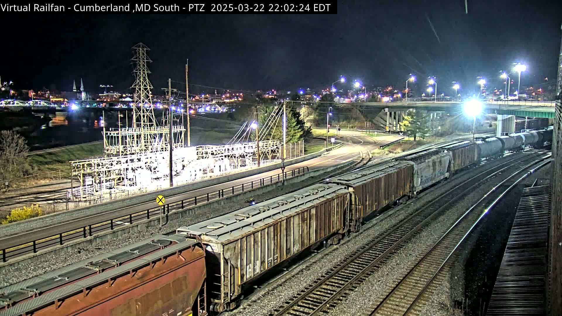 At night, two long freight trains are moving along parallel train tracks near a highway and power substation, under clear skies.