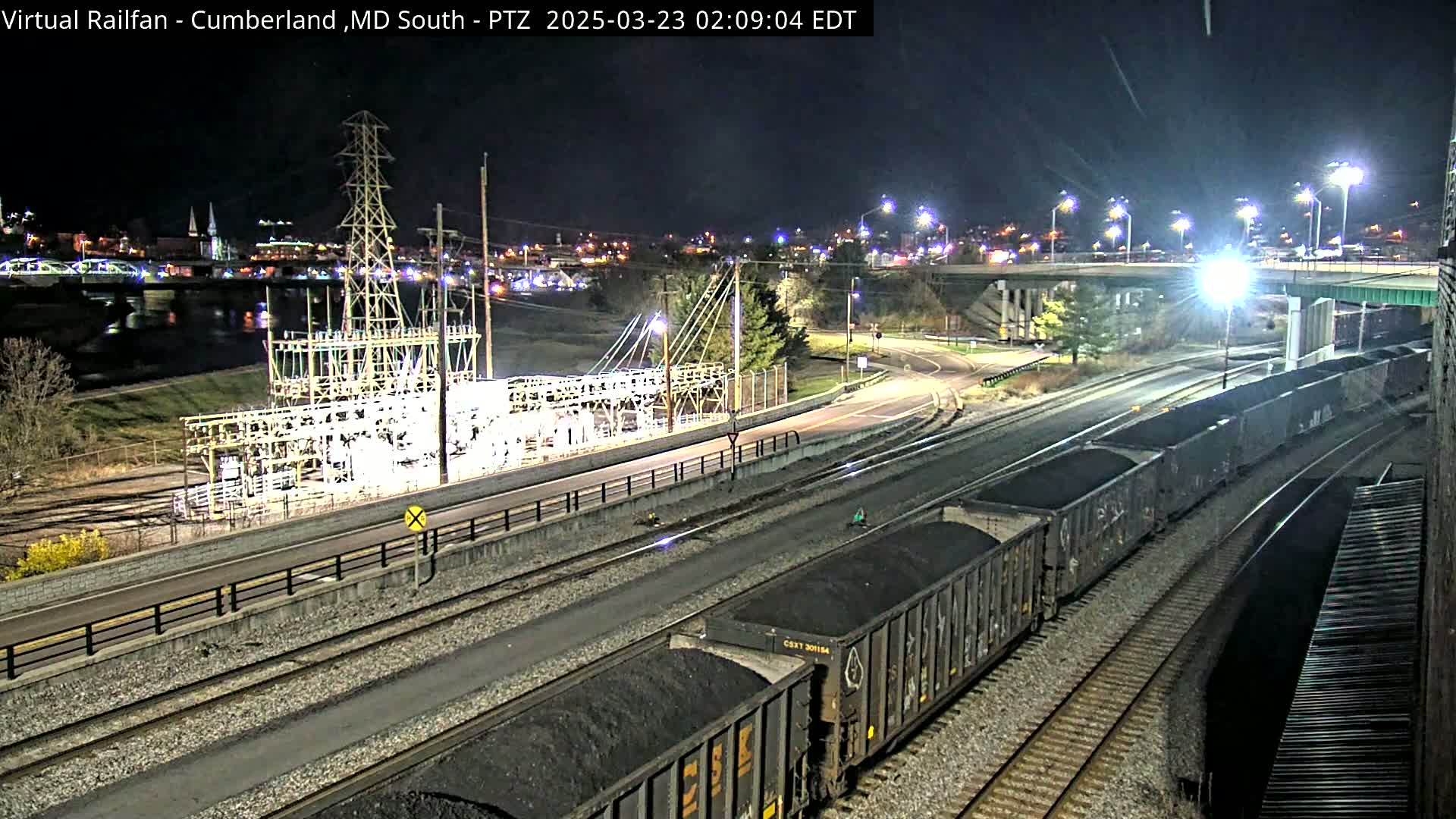 A nighttime view of a railway with coal cars, a road, and an electrical substation near a river under a clear sky.