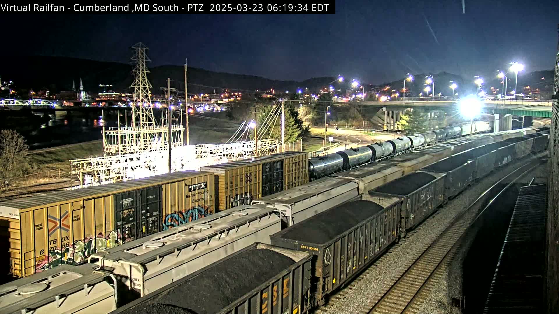 A long freight train, carrying both tankers and coal cars, sits on the tracks at night near a power station and a city lit with streetlights.