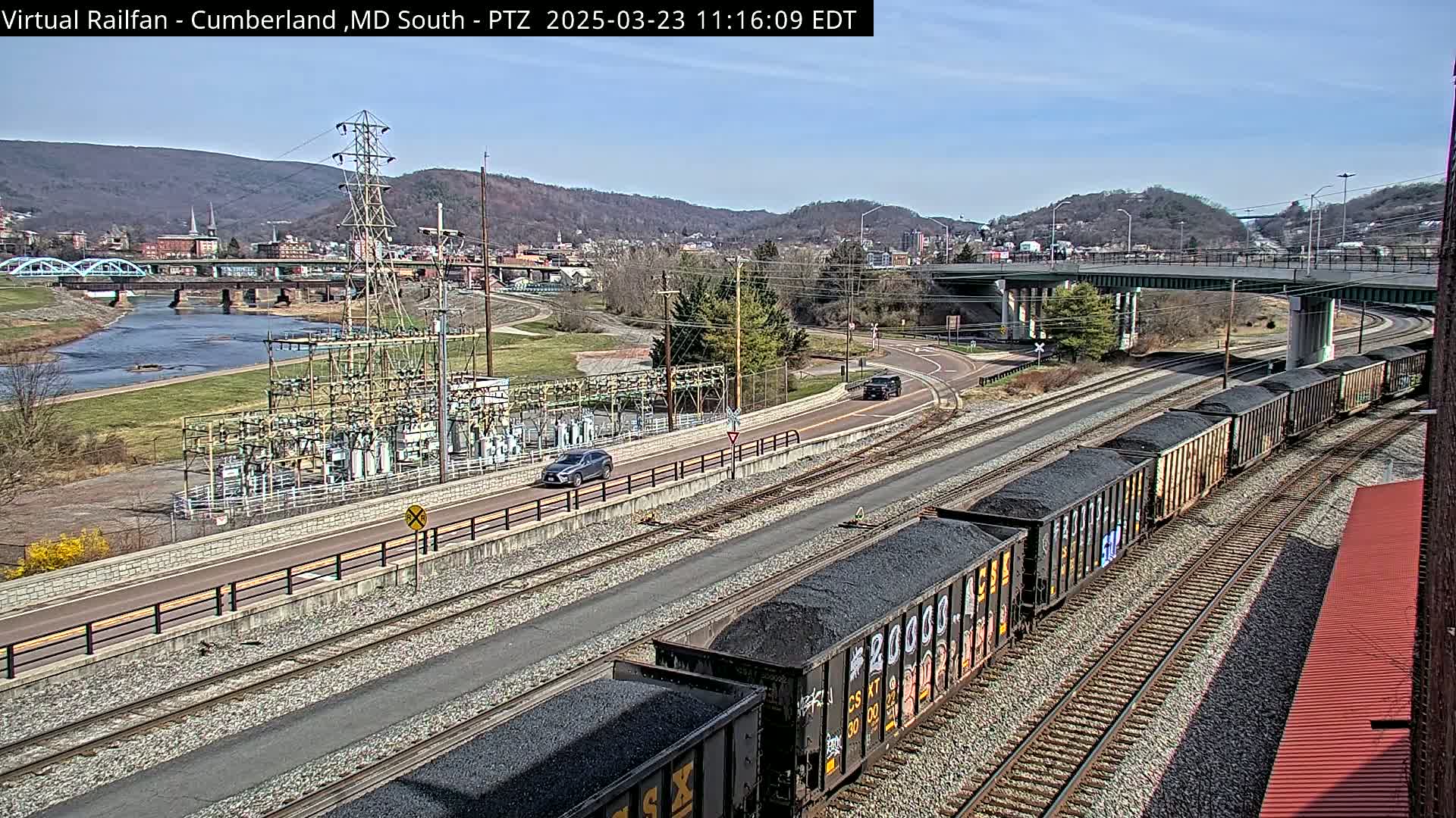 A long train carrying coal is visible on a railway line next to a road and river, under a sunny sky, with a town and mountains in the background.