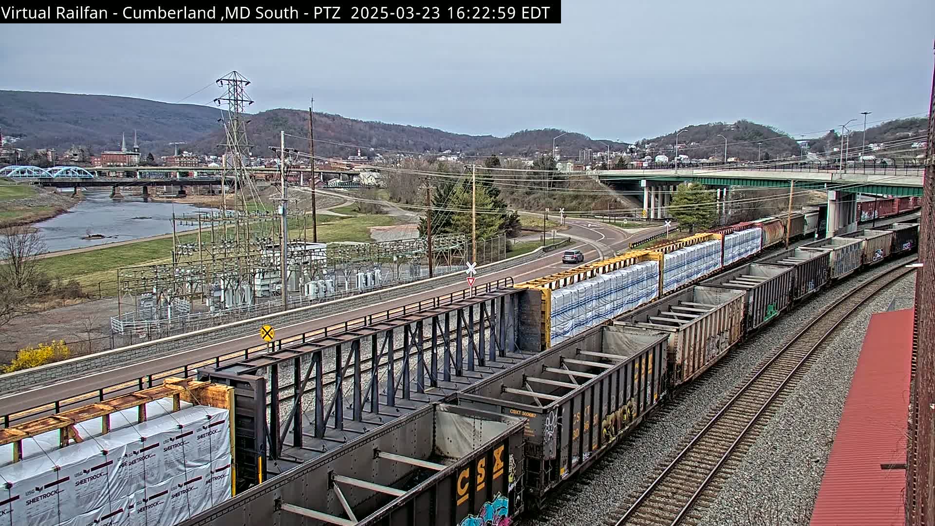 A long freight train carrying packaged materials curves along railroad tracks, passing under a highway overpass, with a river and town visible in the background under an overcast sky.