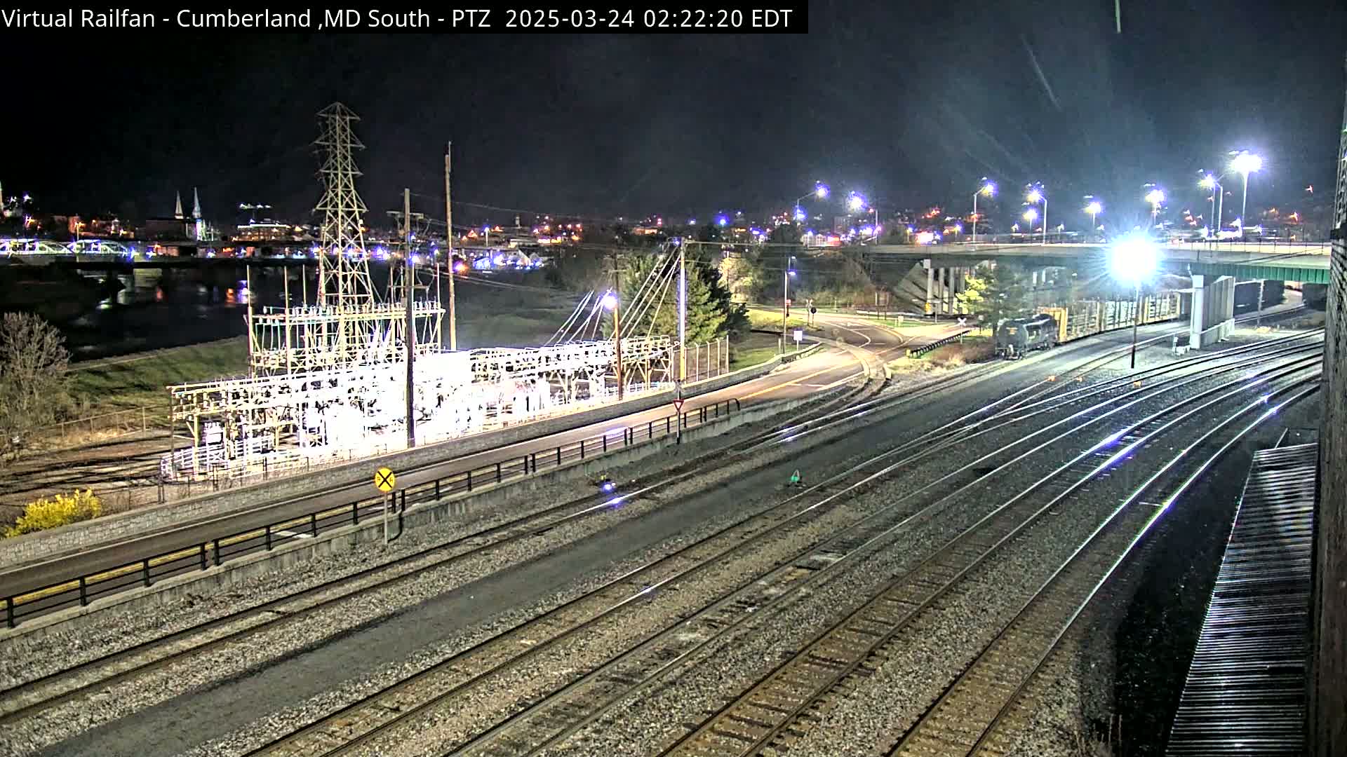 A nighttime view of railroad tracks, a roadway, an electrical substation, a river, and a distant town illuminated by streetlights.