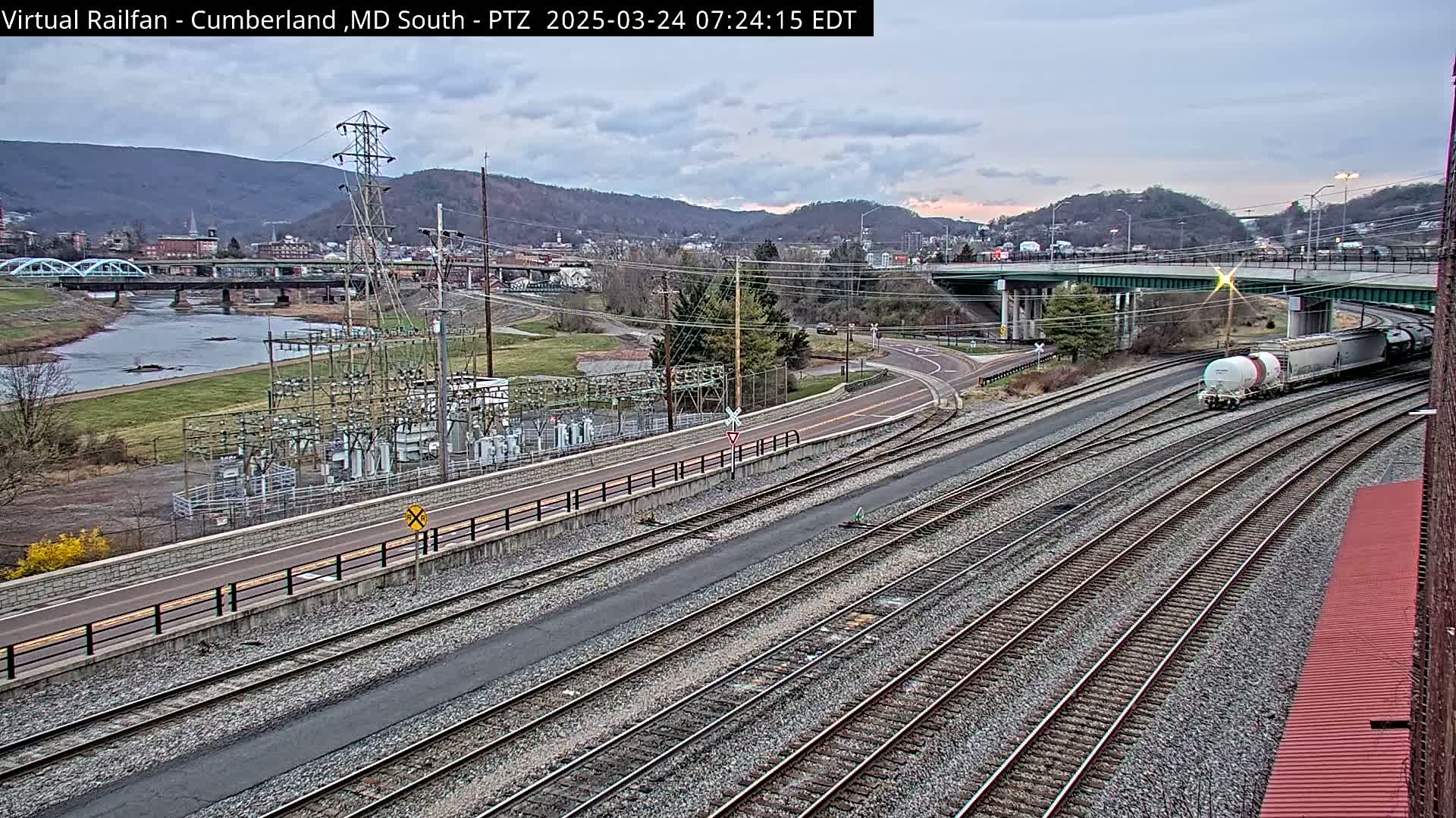 A train passes by a river and a town under an overcast sky, near a power substation and multiple sets of railroad tracks.