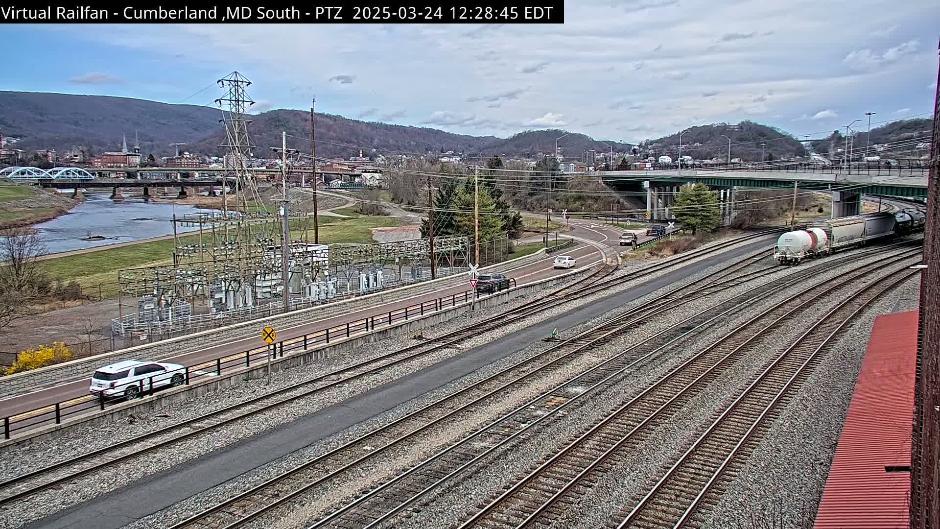 A train passes by a river and a town under a cloudy sky, with multiple sets of railroad tracks in the foreground.