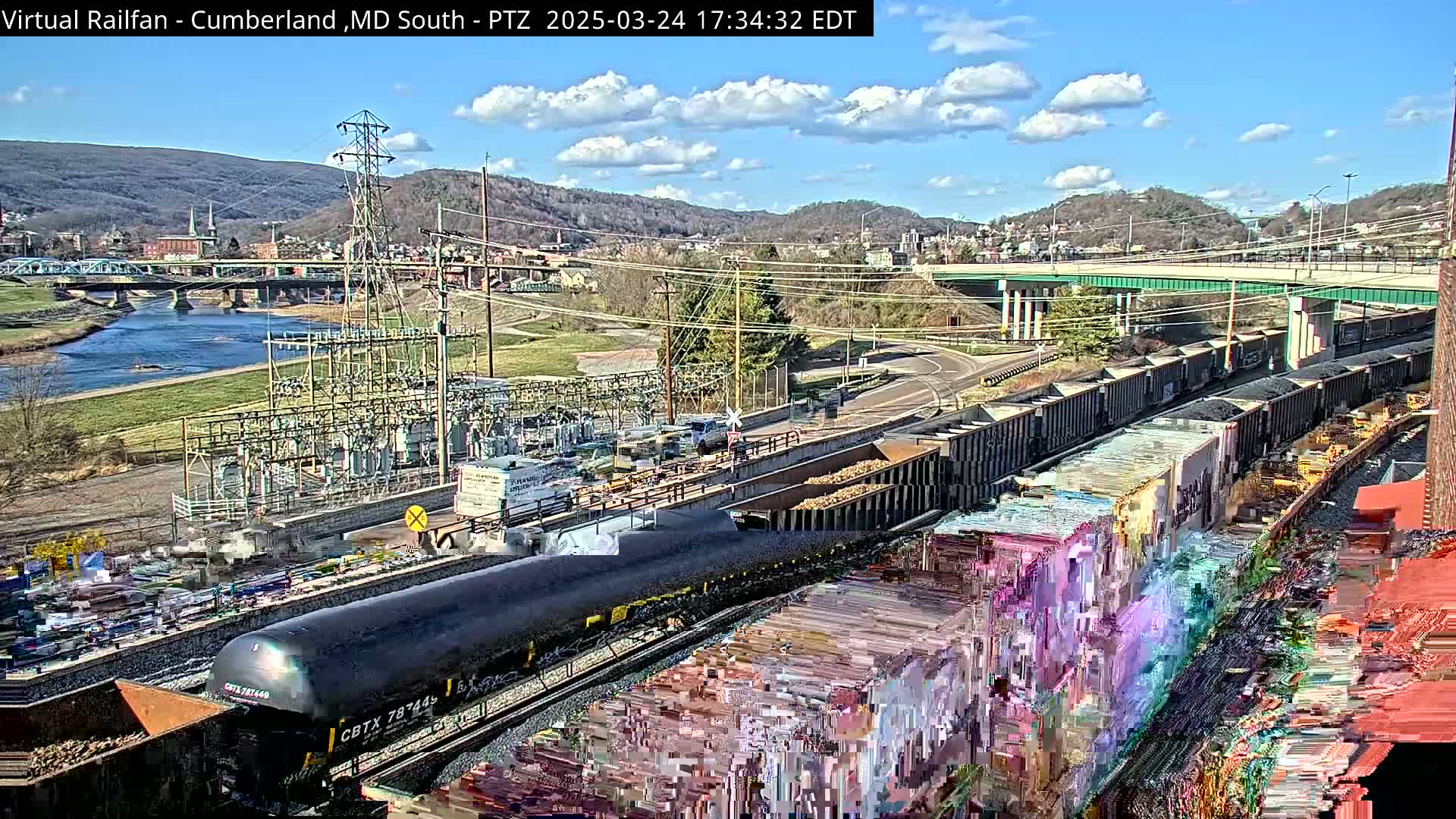 A mostly sunny day reveals a river, a town nestled in hills, and a railway with numerous freight cars, some of which are covered in graffiti.