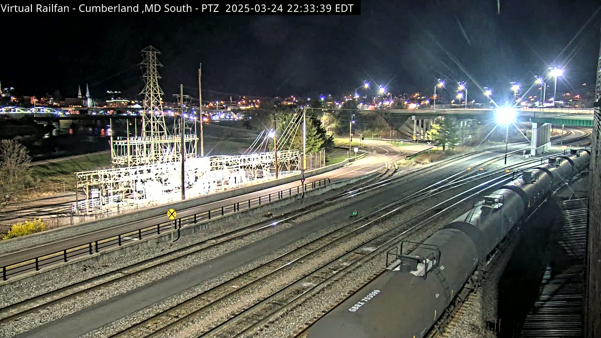 A long freight train of tanker cars is visible at night, passing by an electrical substation near a river and road, under bright artificial lights.