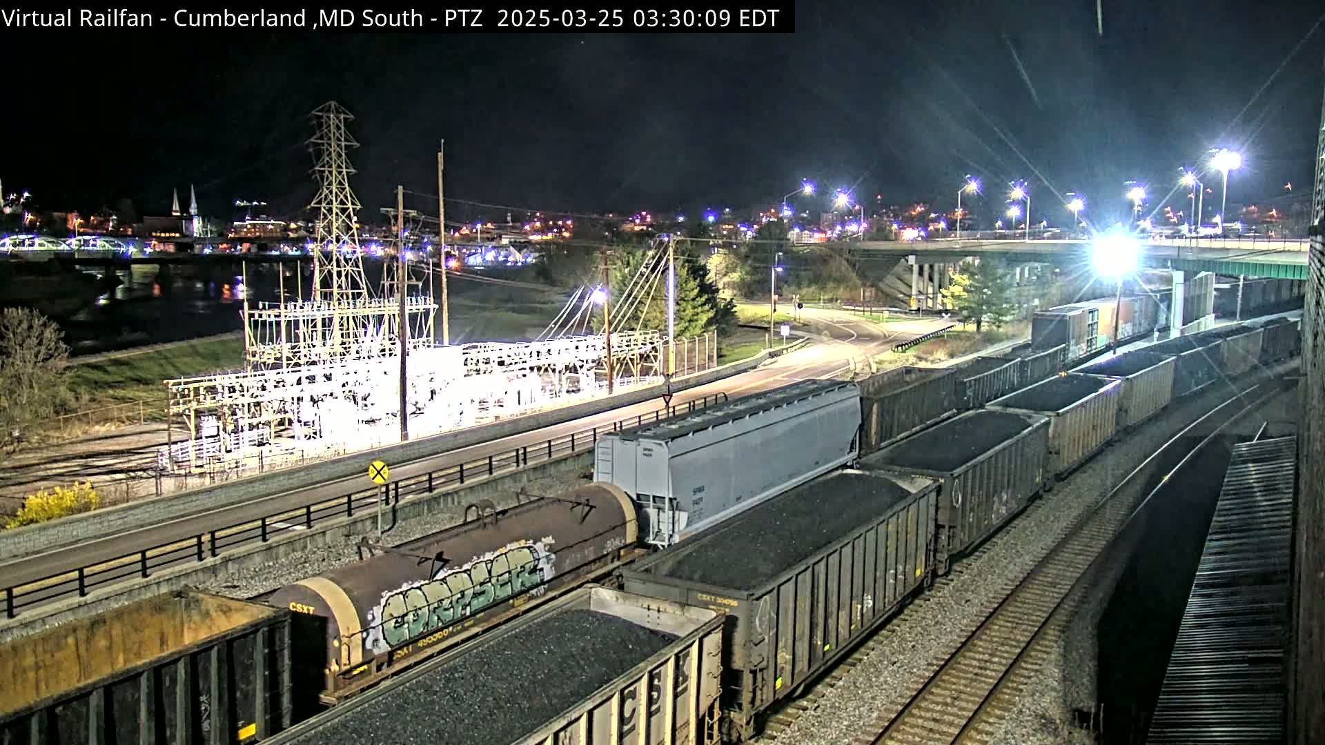 A long freight train carrying coal is parked at night next to a power substation near a river and a highway overpass.