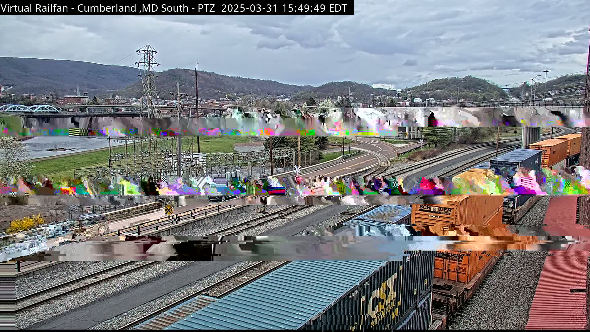 A train with orange and blue shipping containers is passing by a river and a bridge under an overcast sky.