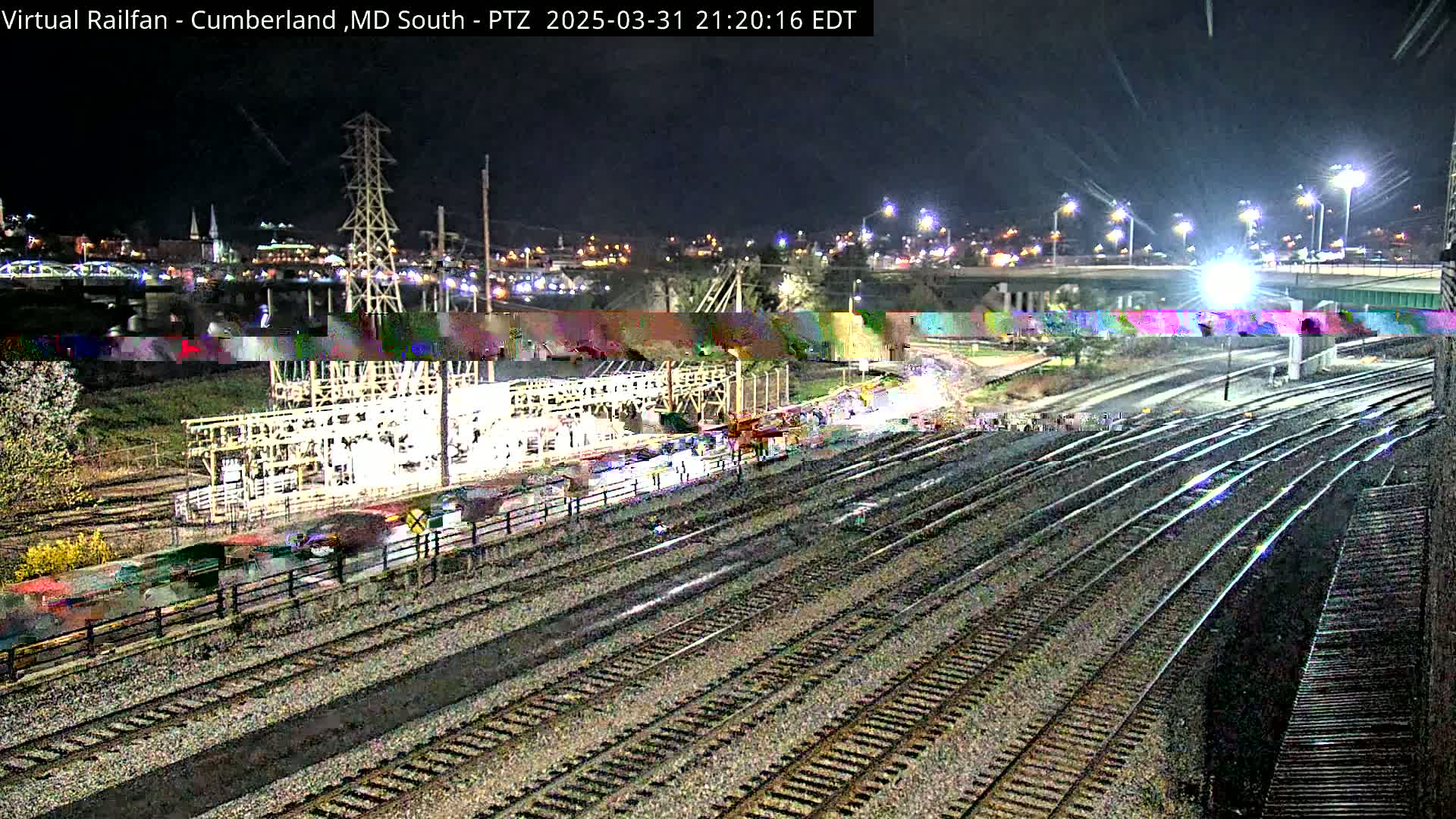 A nighttime view of a railway yard with multiple tracks, a nearby industrial structure, and a city skyline in the background under clear skies.