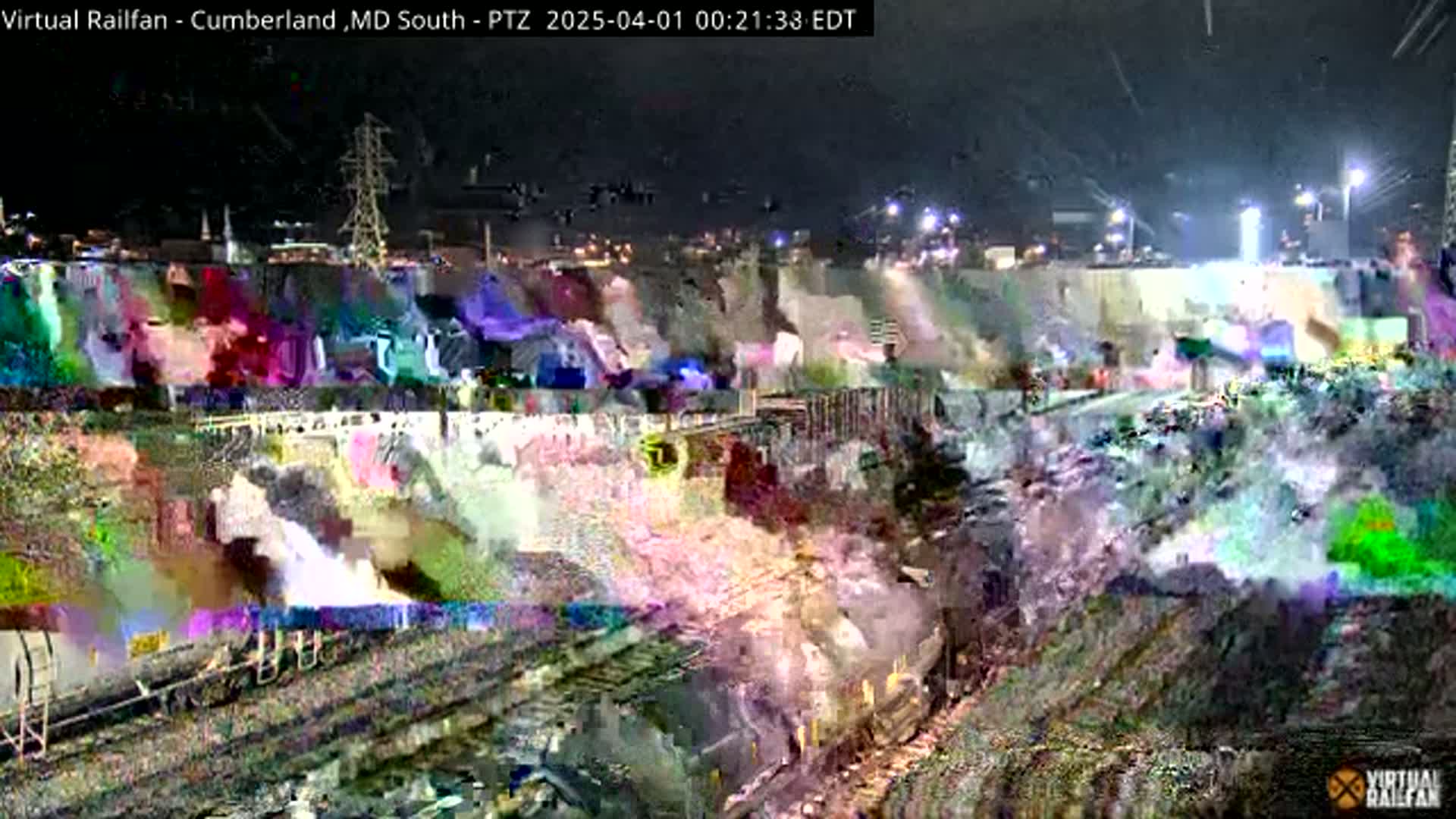 A nighttime, blurry view of a train yard with a large, brightly lit wall in the background.