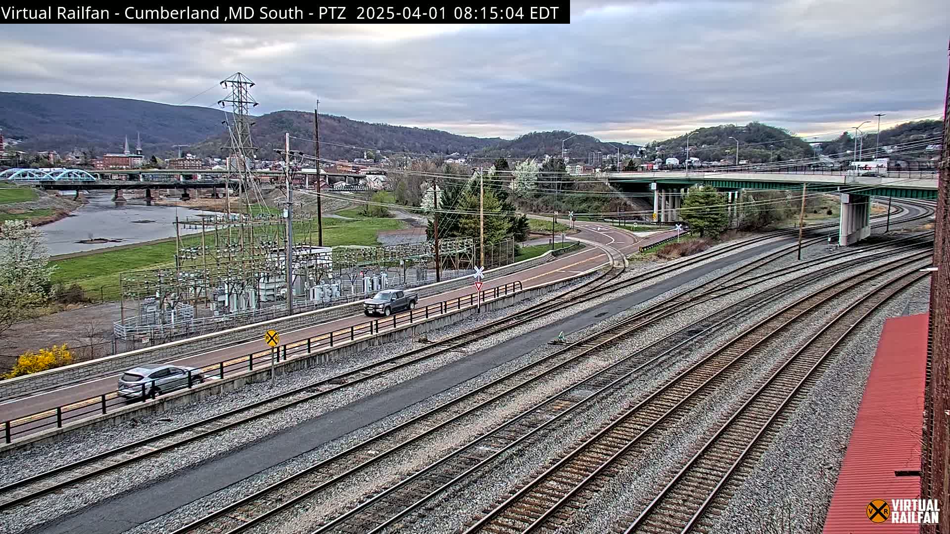 A high-angle, long shot shows several sets of train tracks curving through a landscape with a river, a bridge, a roadway, and some vehicles under an overcast sky.