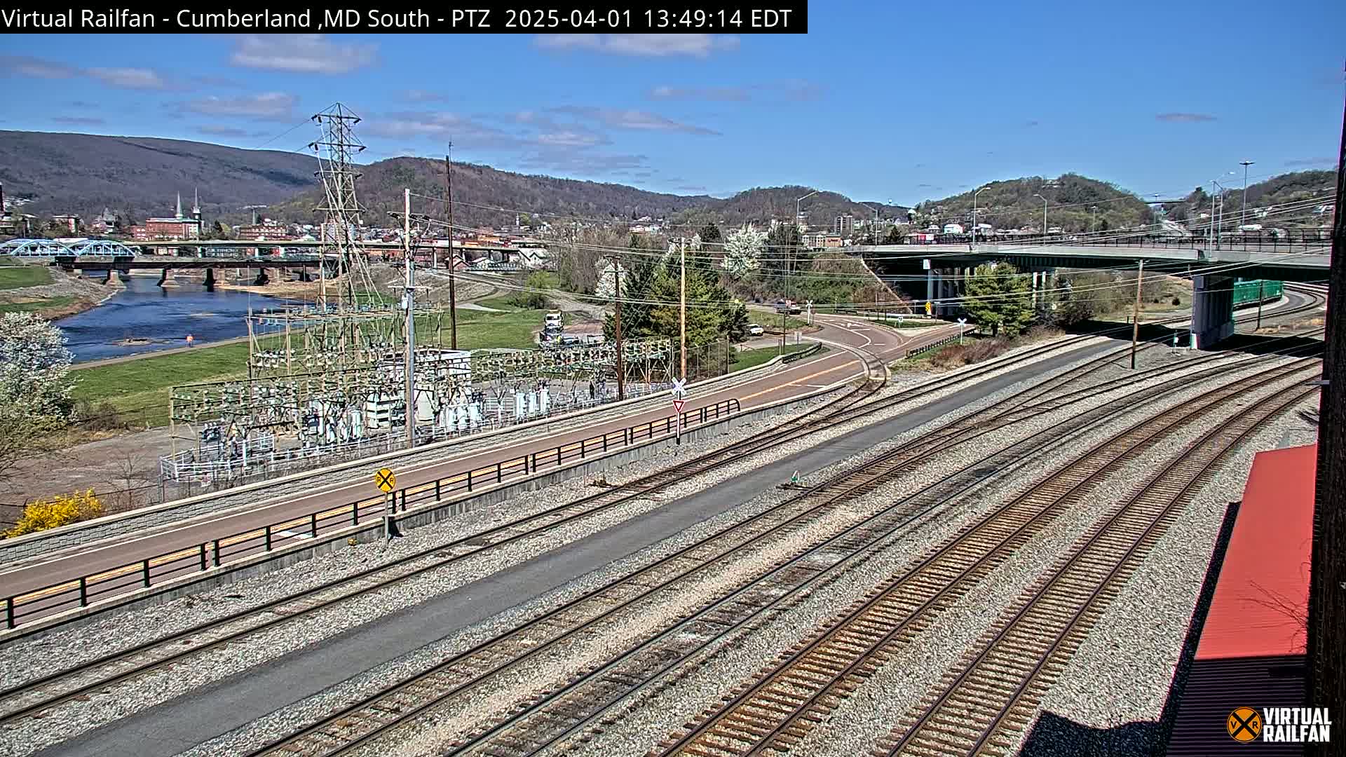 A sunny day reveals a view of railroad tracks leading toward a town nestled in a valley, featuring a river, bridges, and electrical infrastructure.