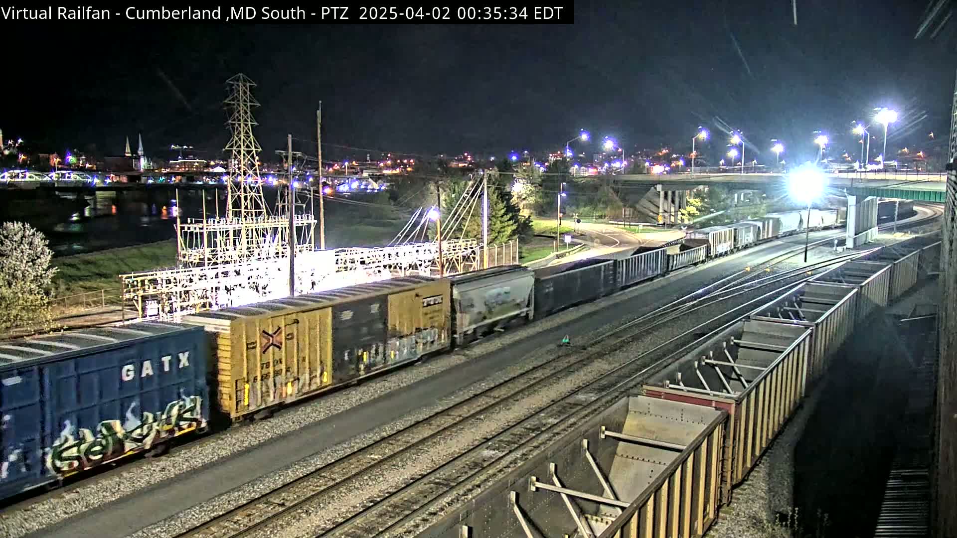 A long freight train travels at night along railroad tracks near a river and city under bright streetlights.