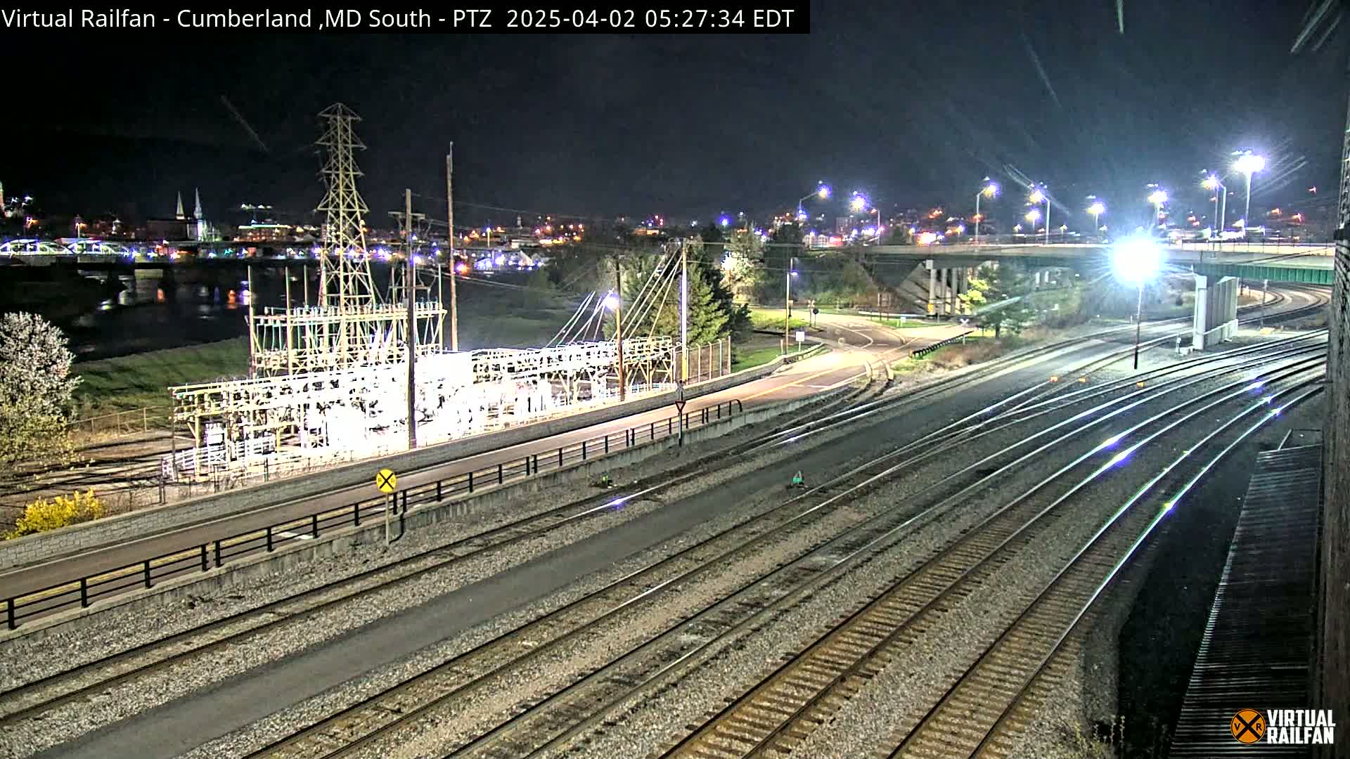 A nighttime view of railroad tracks and roads intersecting near a power substation, with a river and city lights in the background.