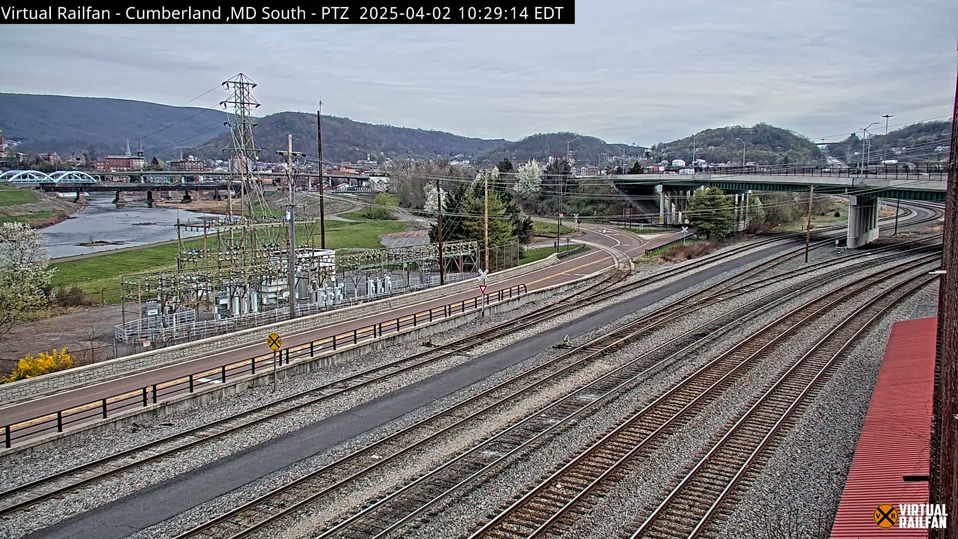 A view of a river, train tracks, a power substation, and a highway overpass on an overcast day, with hills in the background.