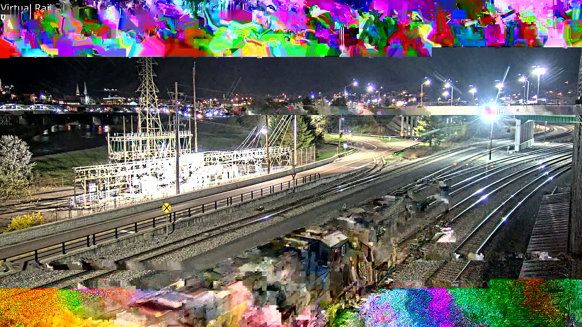 A nighttime view of a train yard and power substation next to a river, with a city skyline in the background under a clear sky.