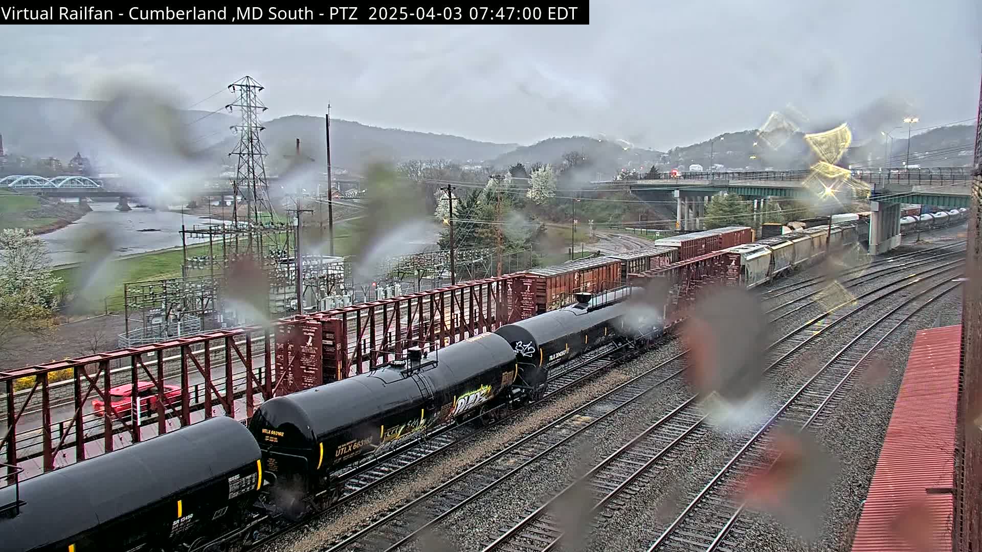 A long freight train, including several tanker cars, sits on a railway line in the rain, with a river and bridge visible in the background.