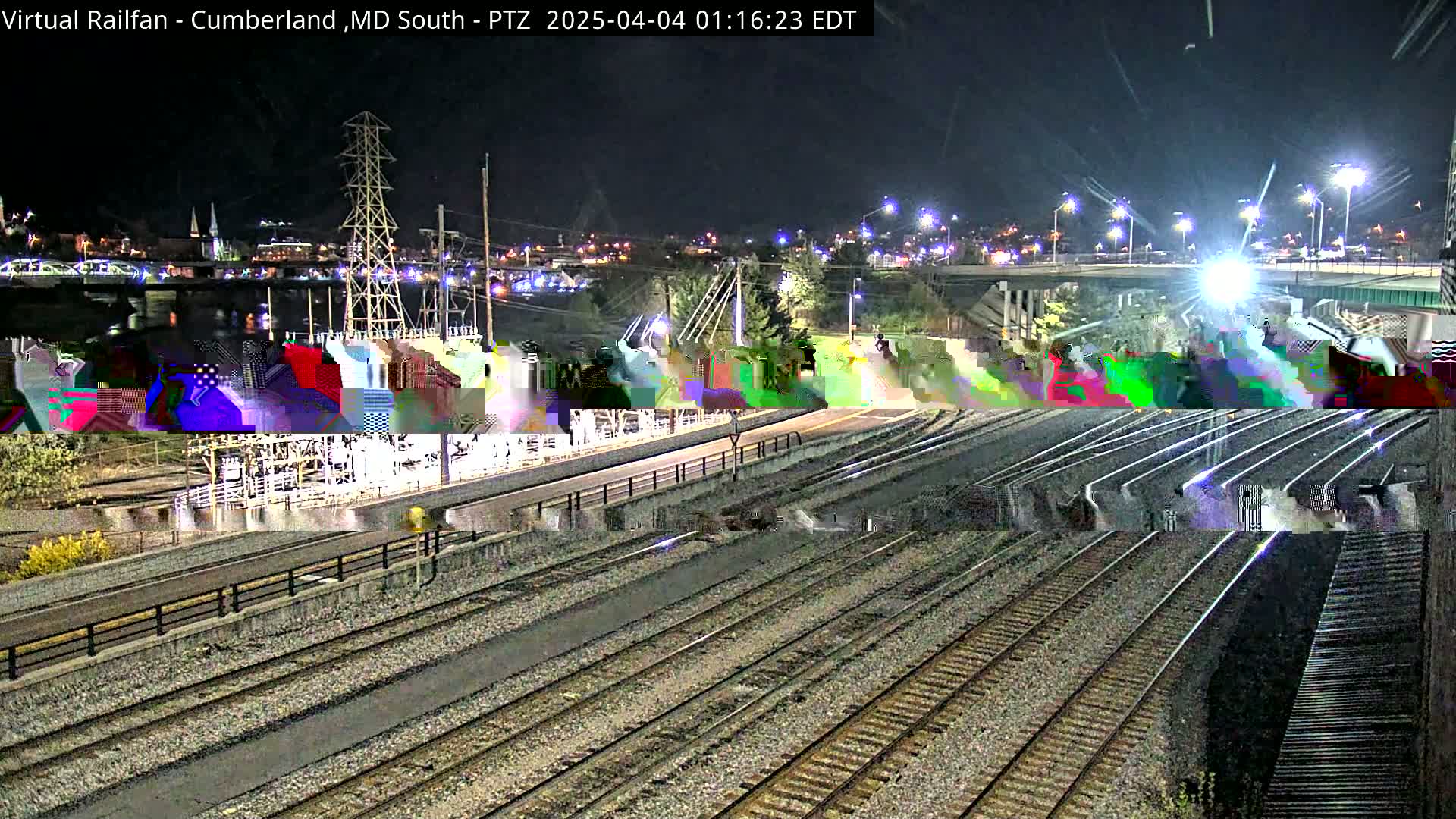 A nighttime view of a rail yard with multiple train tracks, a nearby river, and a city skyline in the distance.