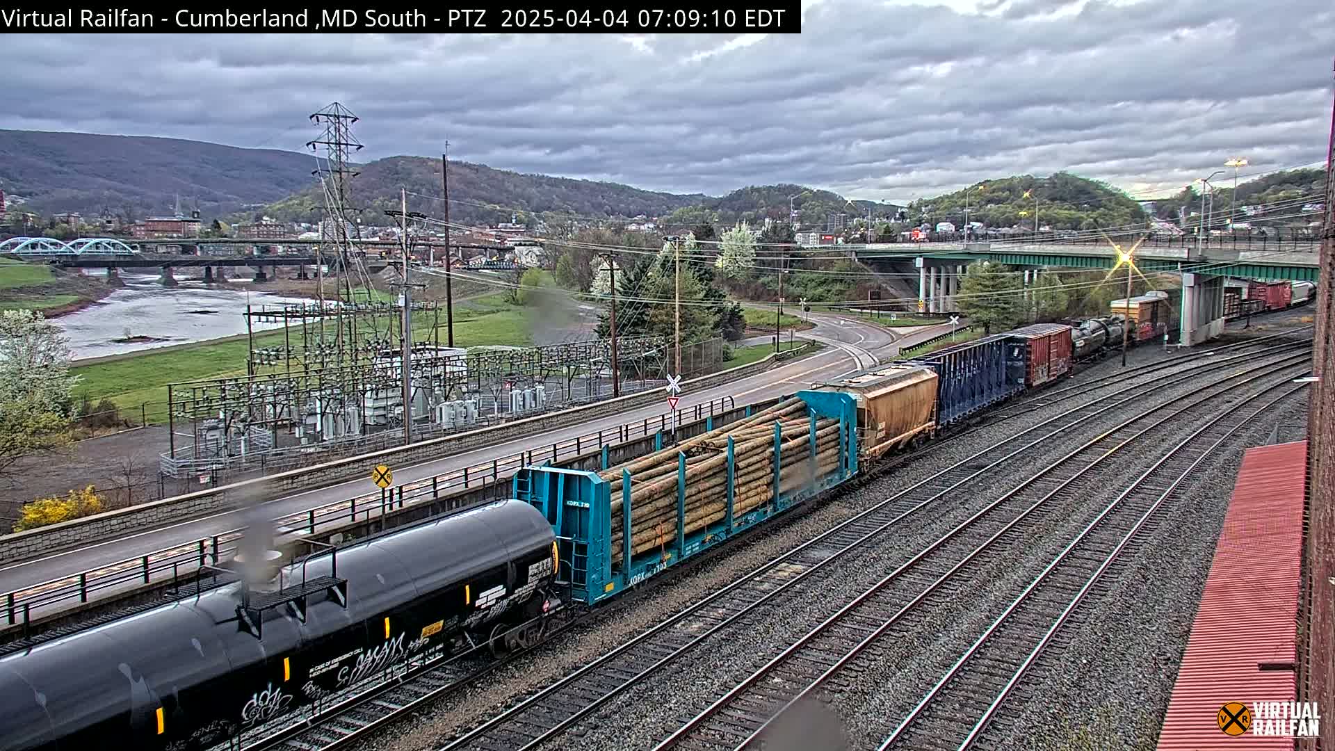 A freight train carrying logs and tanker cars is visible on railway tracks next to a river and a town under a cloudy sky.