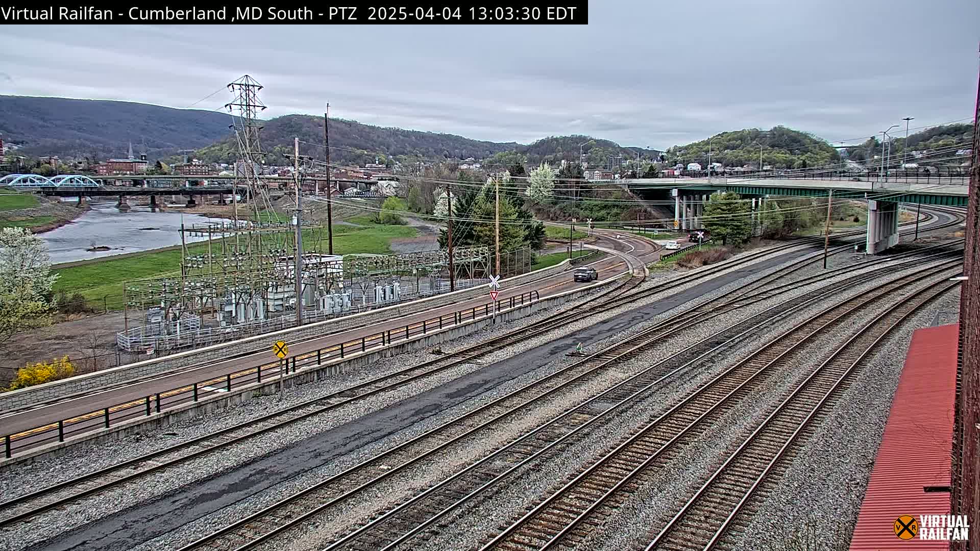 A high-angle, long shot shows a railroad yard with multiple train tracks, a river, a bridge, and a town under an overcast sky.