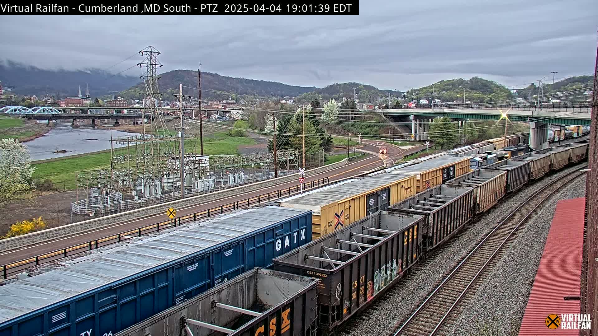 A long freight train curves along railroad tracks under an overcast sky, passing by a river, bridge, and power substation.