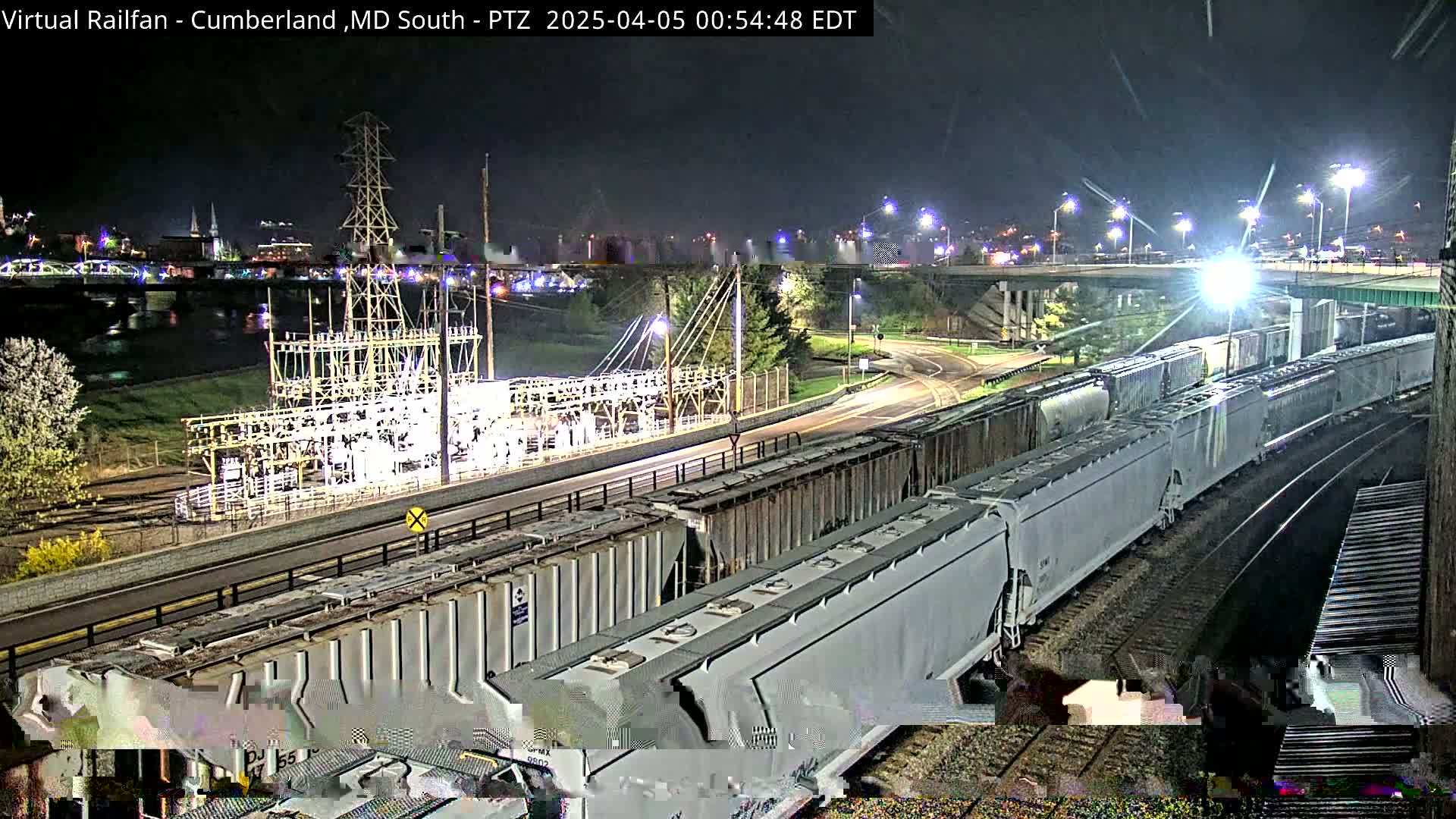 A long freight train is moving through a nighttime scene, near a river, power station, and bridge, under clear skies.