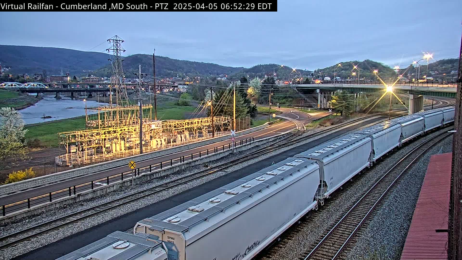 A long freight train curves along railroad tracks next to a river and a power substation under a twilight sky.