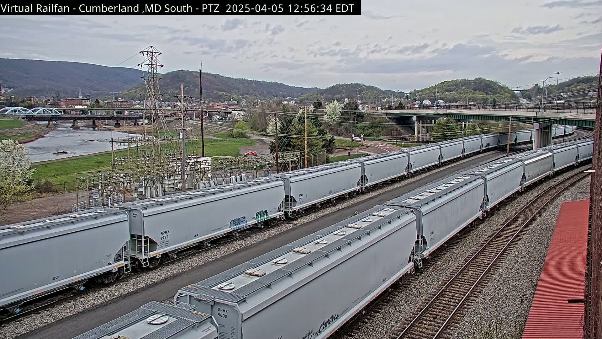 Two long trains of light gray covered hopper cars are parked on parallel railroad tracks beside a river, under an overcast sky, with a town and mountains in the background.